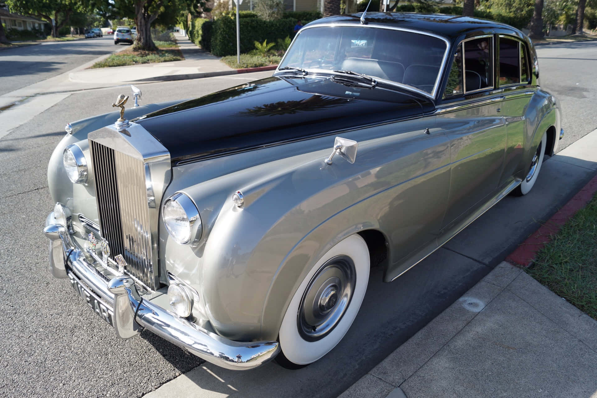 Bentley S2 桌布 壁紙 — 一輛閃亮的銀色勞斯萊斯，黑色車頂，停泊在陽光明媚的郊區街道上，反射著樹木和天空。