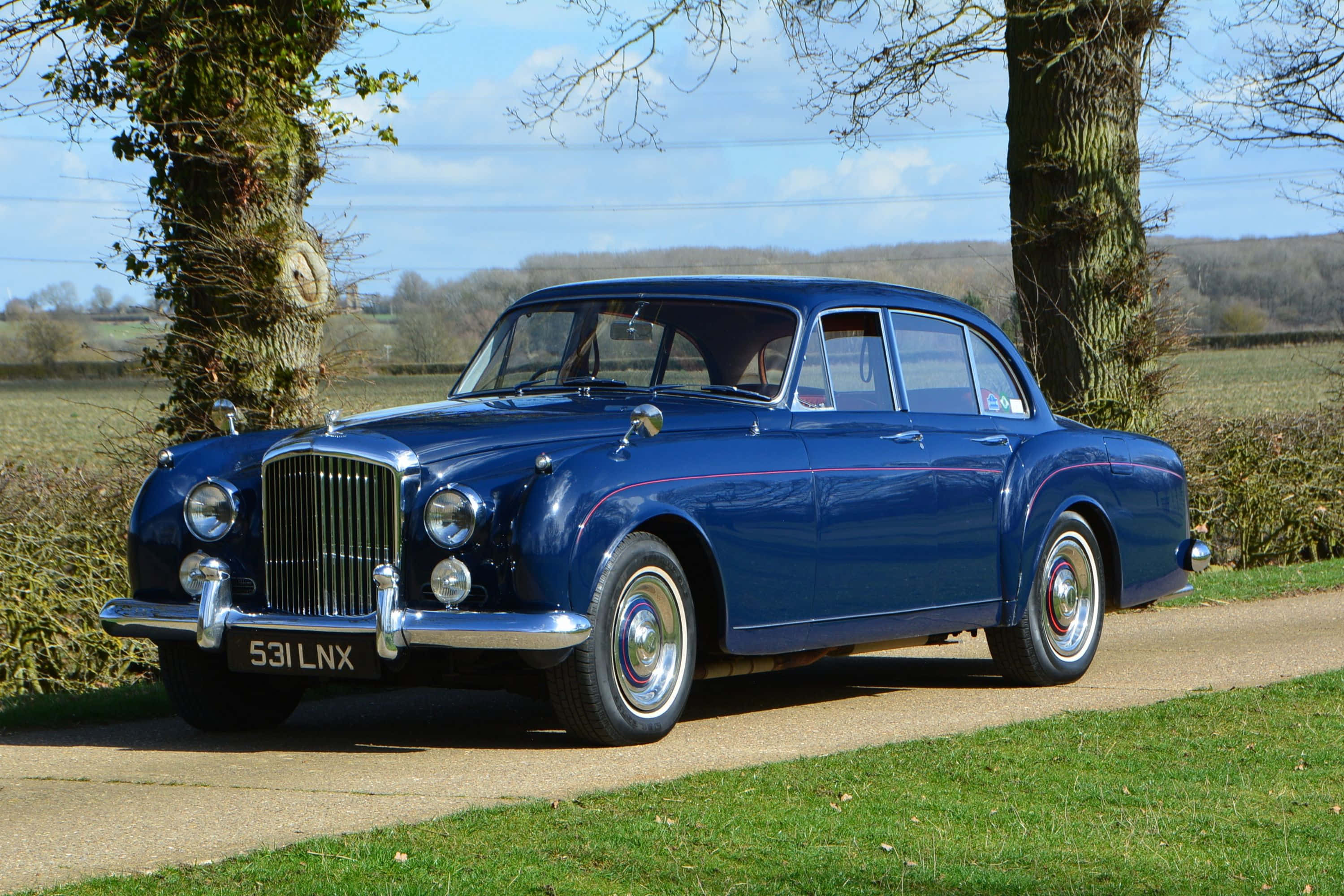 Bentley S2 桌布 壁紙 — 一輛閃亮的藍色經典賓利轎車停泊在碎石車道上，兩旁是無葉的樹木，上方是明亮的藍天。