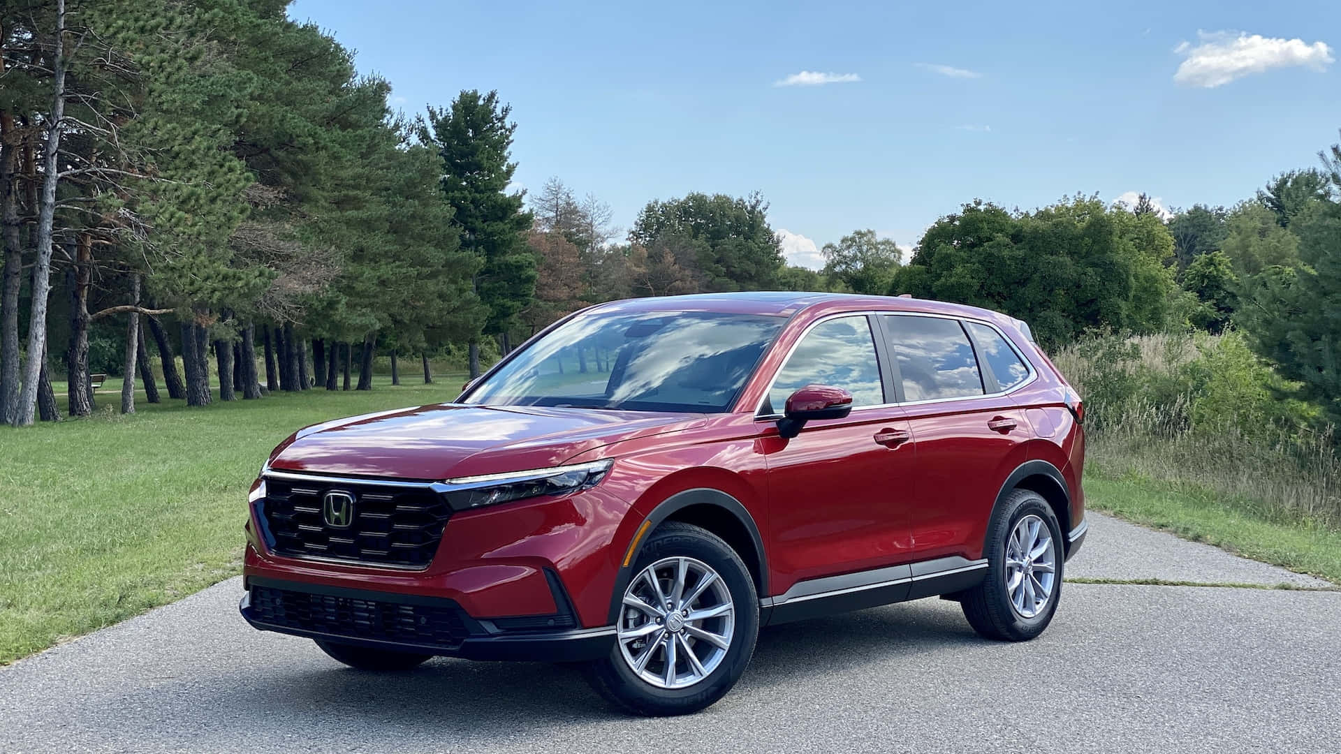 Honda Cr-v 桌布 壁紙 — 一輛亮麗的紅色本田SUV停放在鋪設的道路上，周圍是高聳的樹木和綠草，在晴朗的藍天下。