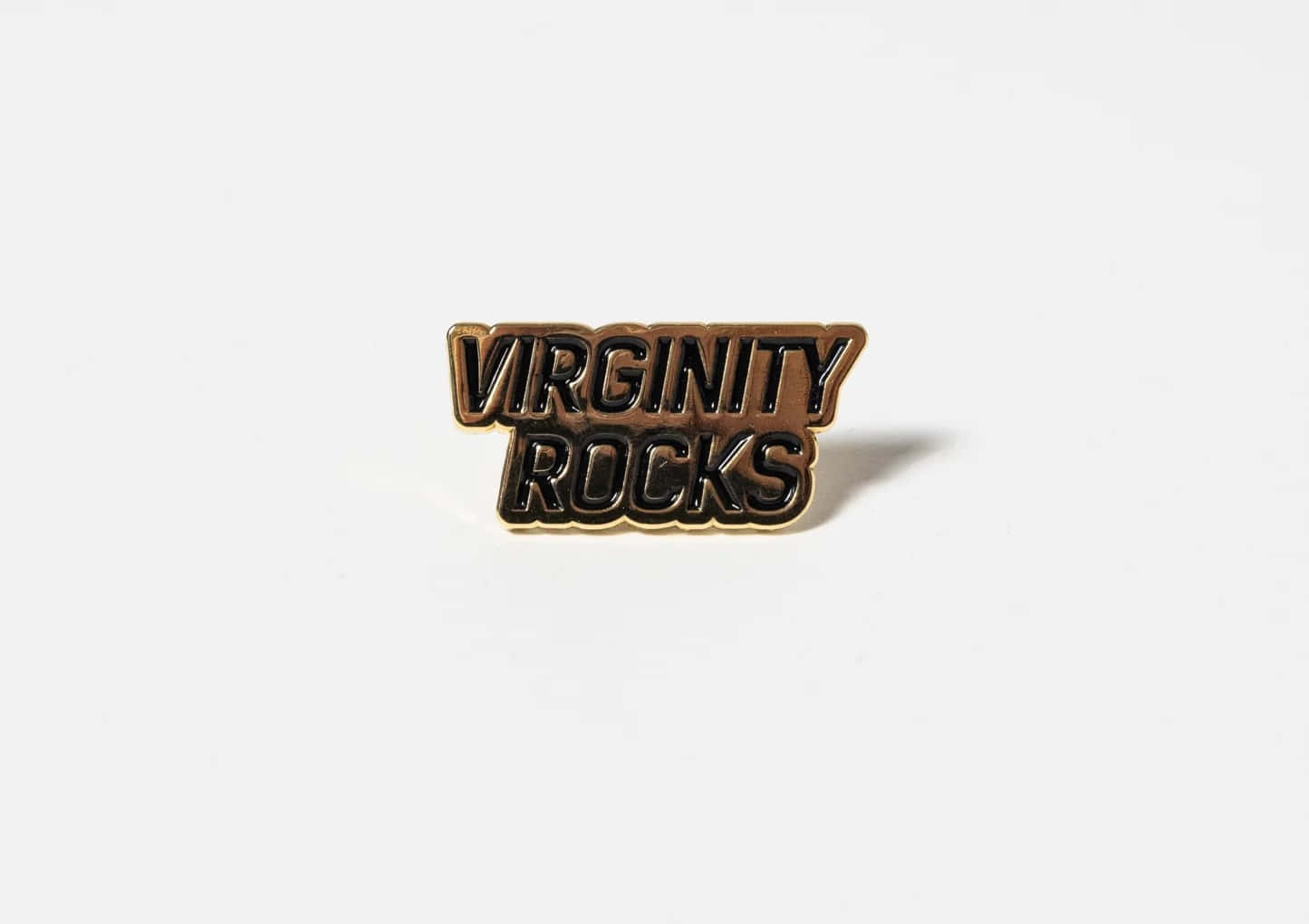  壁紙 — 一枚金色和黑色的琺瑯別針，上面寫著「VIRGINITY ROCKS」，放在純白色的表面上。
