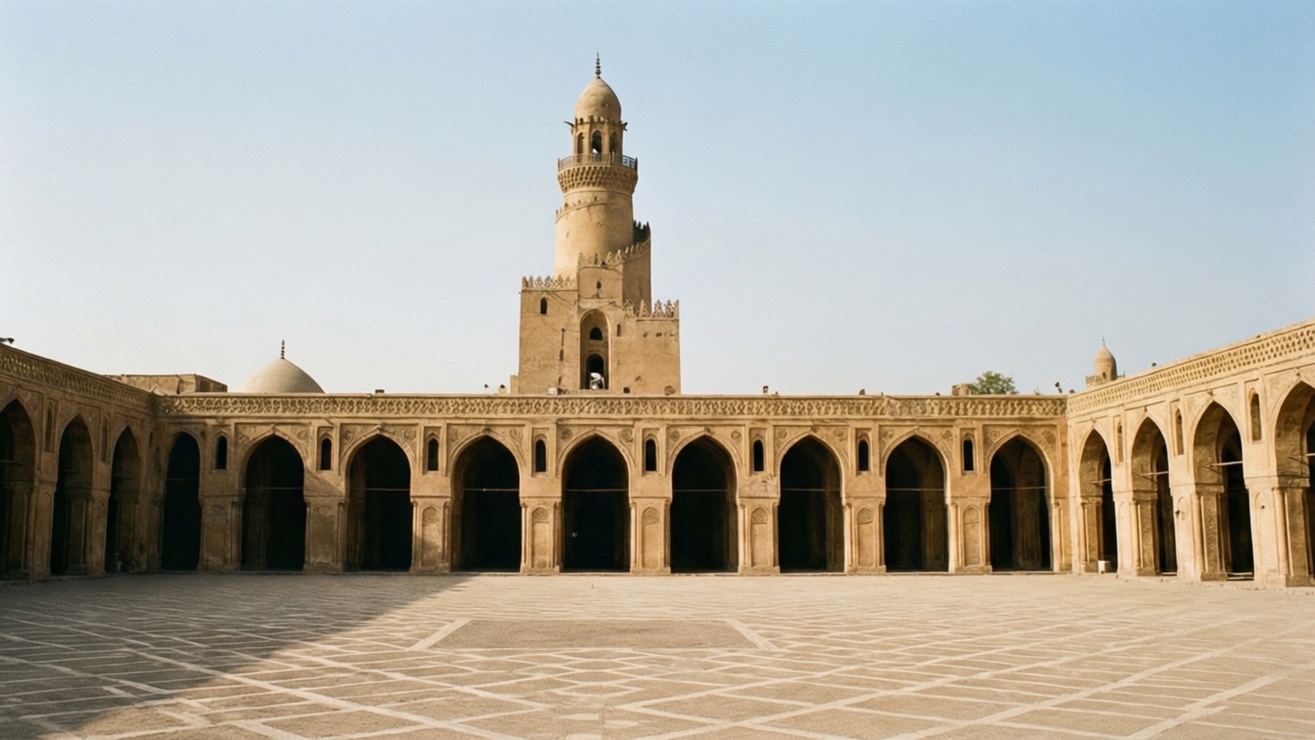 Ibn Tulun Mosque Serenity 壁紙 — 一個寬敞的陽光庭院，擁有拱形柱廊和一座高聳的宣禮塔，在清澈的藍天下。
