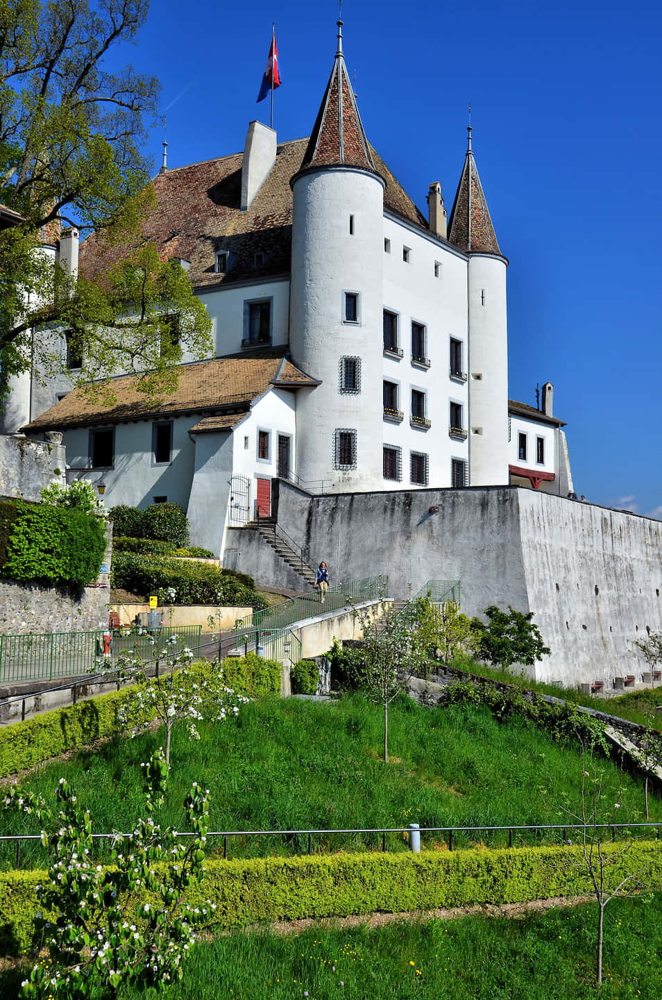 Nyon Switzerland 桌布 壁紙 — 一座宏偉的白色城堡，擁有圓錐形屋頂和石牆，矗立在鬱鬱蔥蔥的綠色山丘上，天空清澈湛藍。