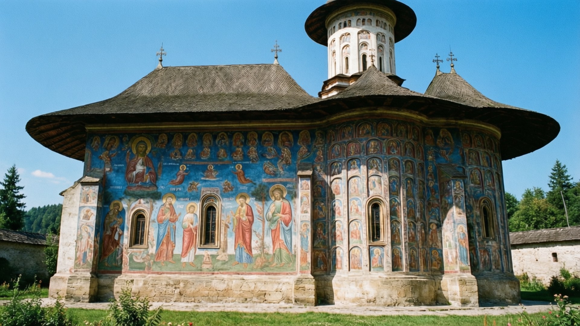 Moldovita Monastery Frescoes 壁紙 — 一座大型木造教堂，藍色牆壁上佈滿宗教壁畫，矗立在晴朗的藍天下，周圍環繞著綠樹。
