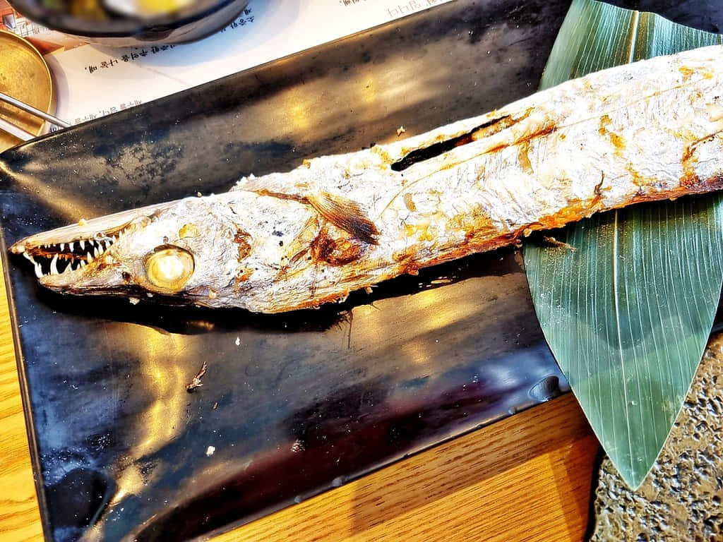  壁紙 — 一條烤魚，有尖銳的牙齒和金棕色魚皮，放在黑色盤子上，旁邊有一片綠葉。