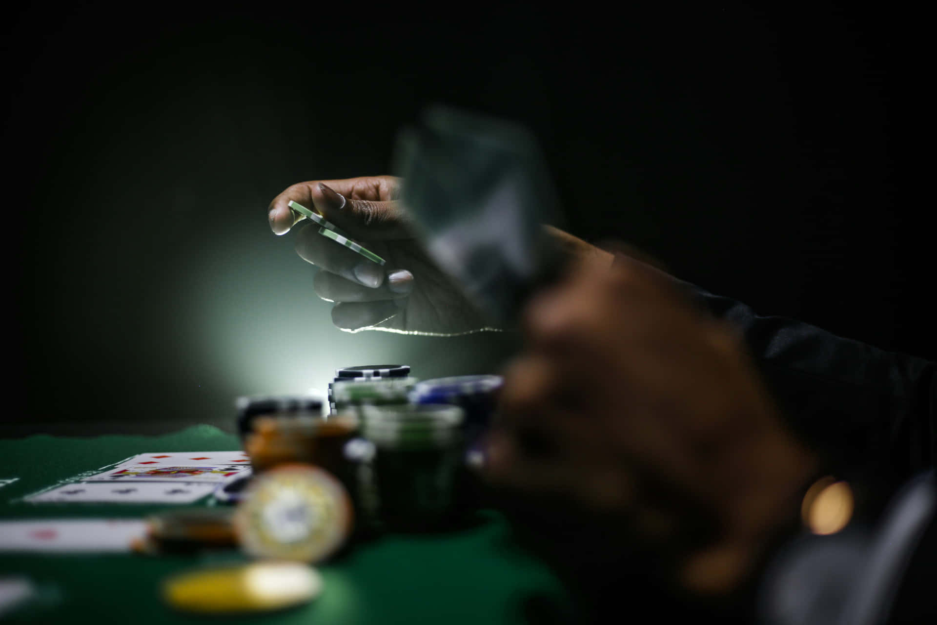  壁紙 — 在黑暗中，一隻手在戲劇性的聚光燈下，拿著一張撲克牌，放在綠色毛氈桌上，桌上有籌碼。