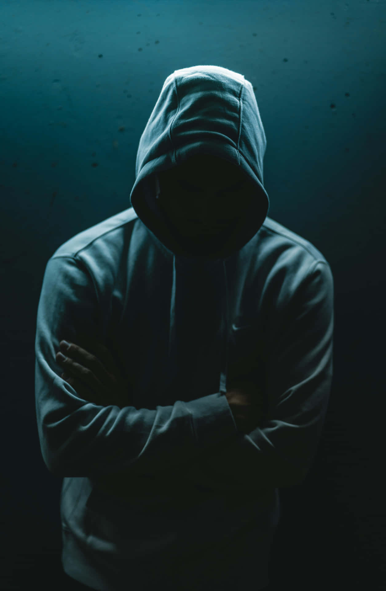  壁紙 — 一個戴著連帽衫的人物交叉雙臂站在昏暗的藍光中，臉部隱藏在陰影中，背景是深色紋理牆壁。