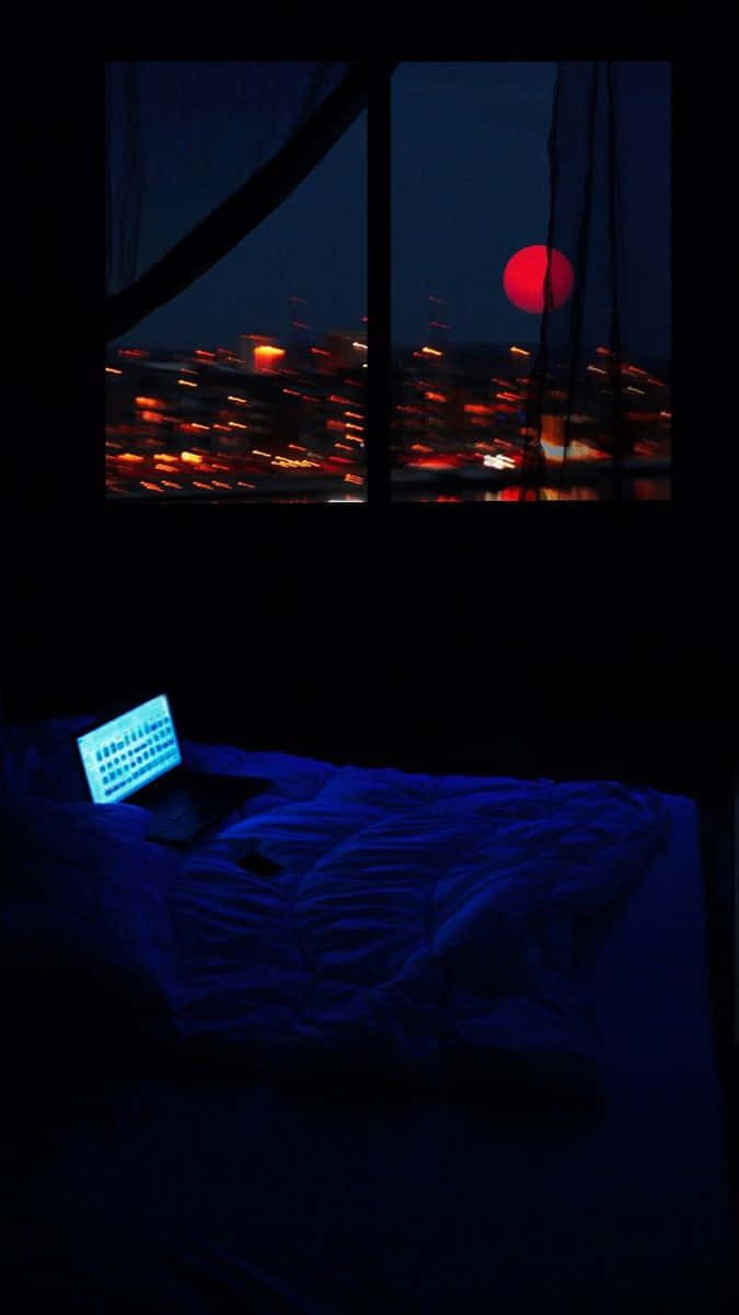 HD Aesthetic 桌布 壁紙 — 一台筆記型電腦在床上發光，窗戶下顯示紅色月亮和模糊的城市燈光在夜晚。