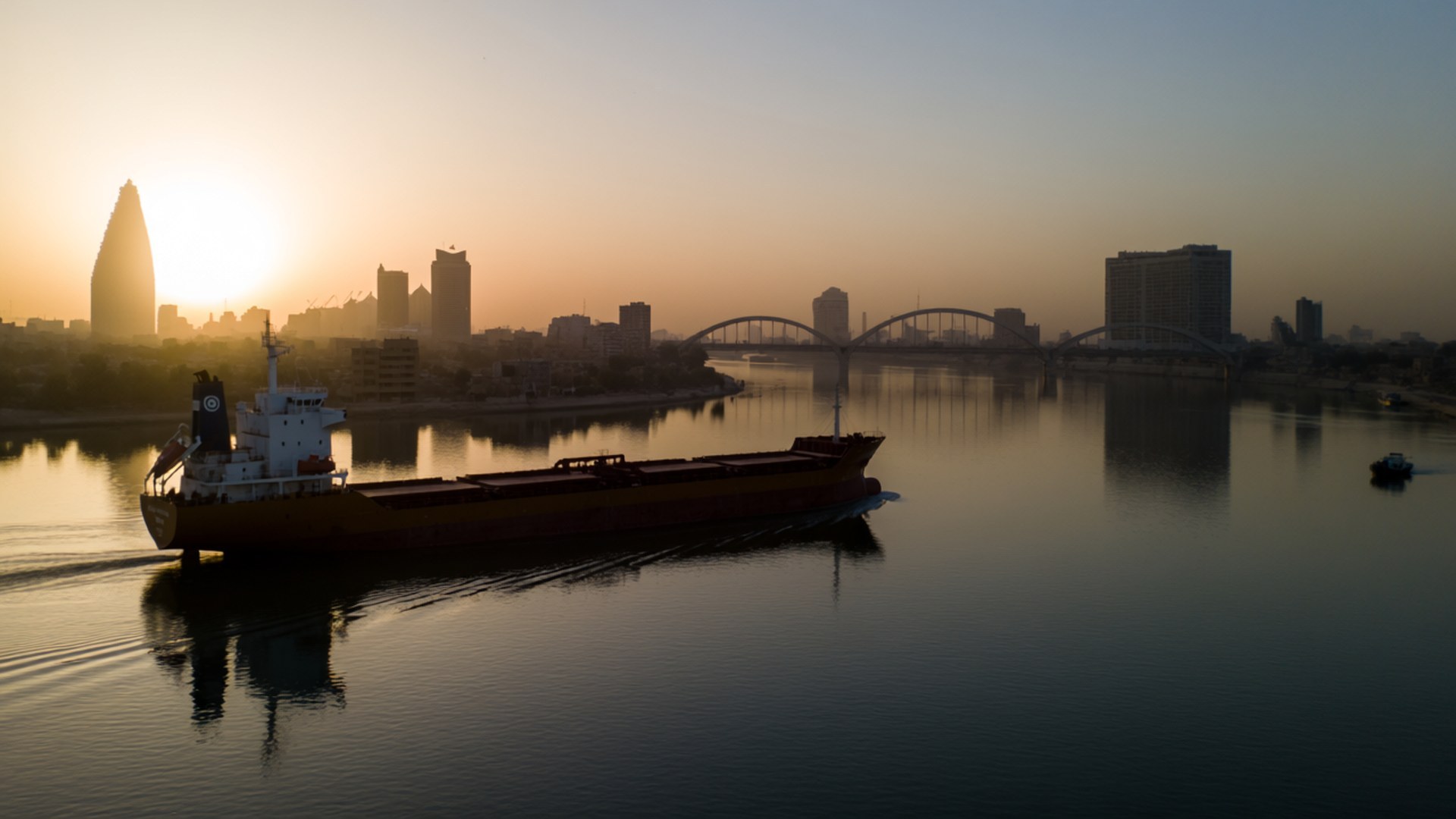 Tigris River Sunset 壁紙 — 一艘大型貨船在日出時平靜地航行在寬闊的河流上，背景朦朧中可見現代城市天際線和拱橋。
