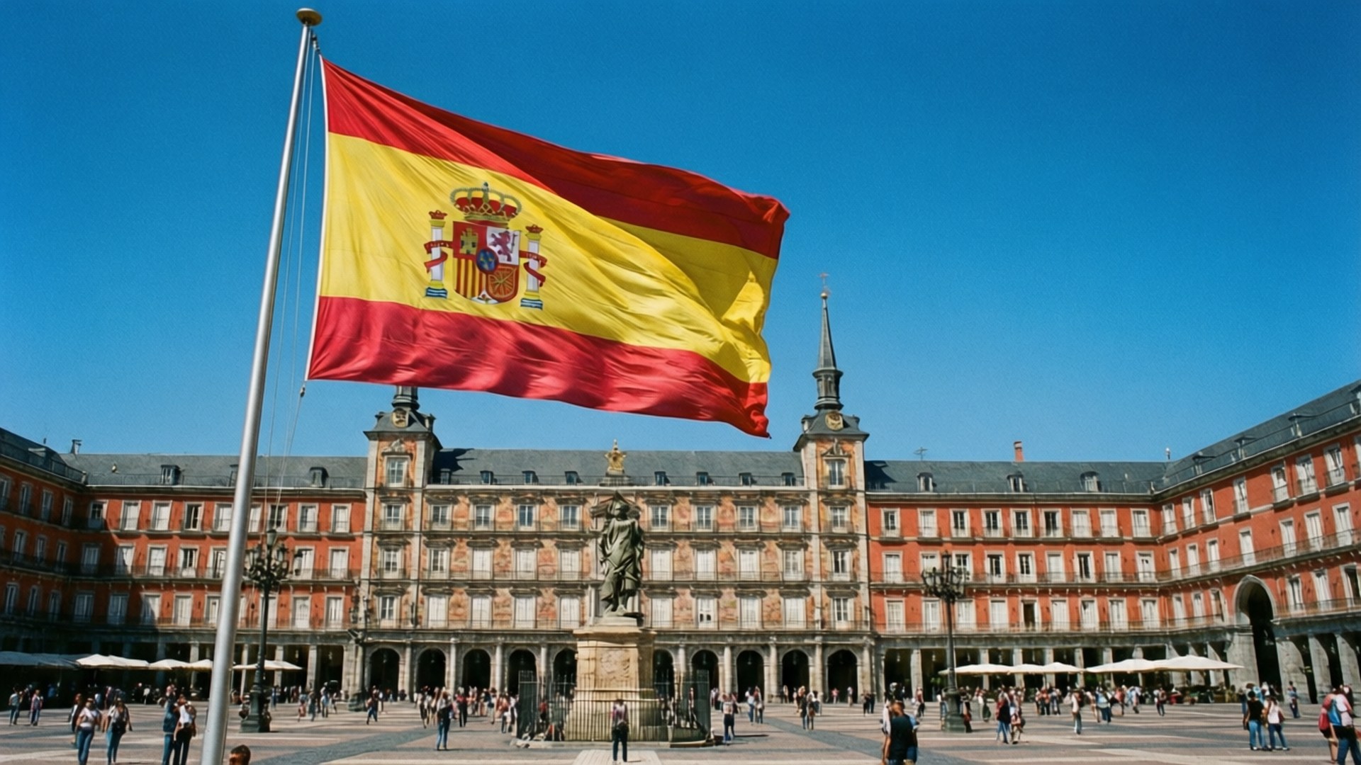 Madrid Plaza Flag 壁紙 — 一面大型西班牙國旗在馬德里歷史悠久的馬約爾廣場前，在晴朗的藍天下自豪地飄揚。