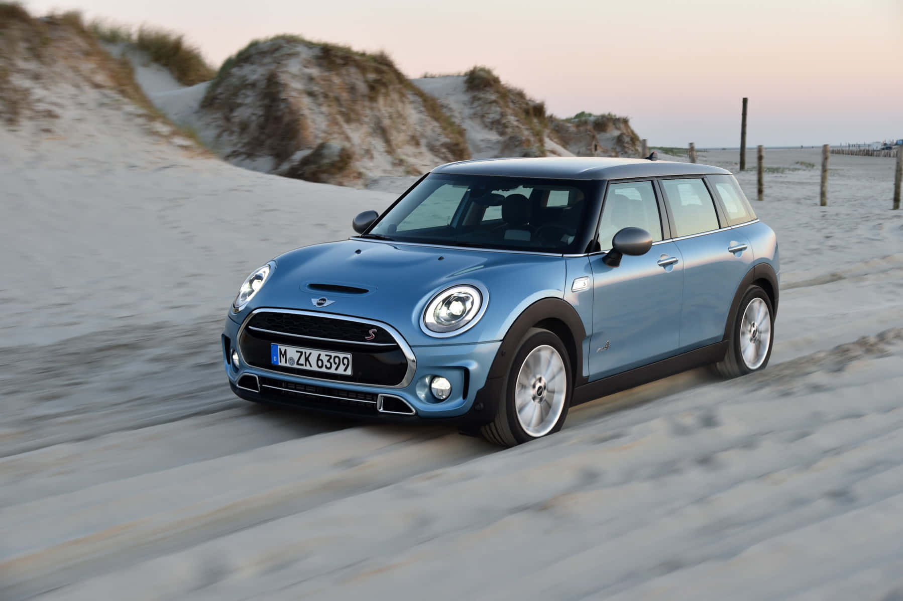 Mini Cooper S Clubman All4 桌布 壁紙 — 一輛淺藍色MINI Countryman在日落時分快速駛過沙灘，背景有沙丘。