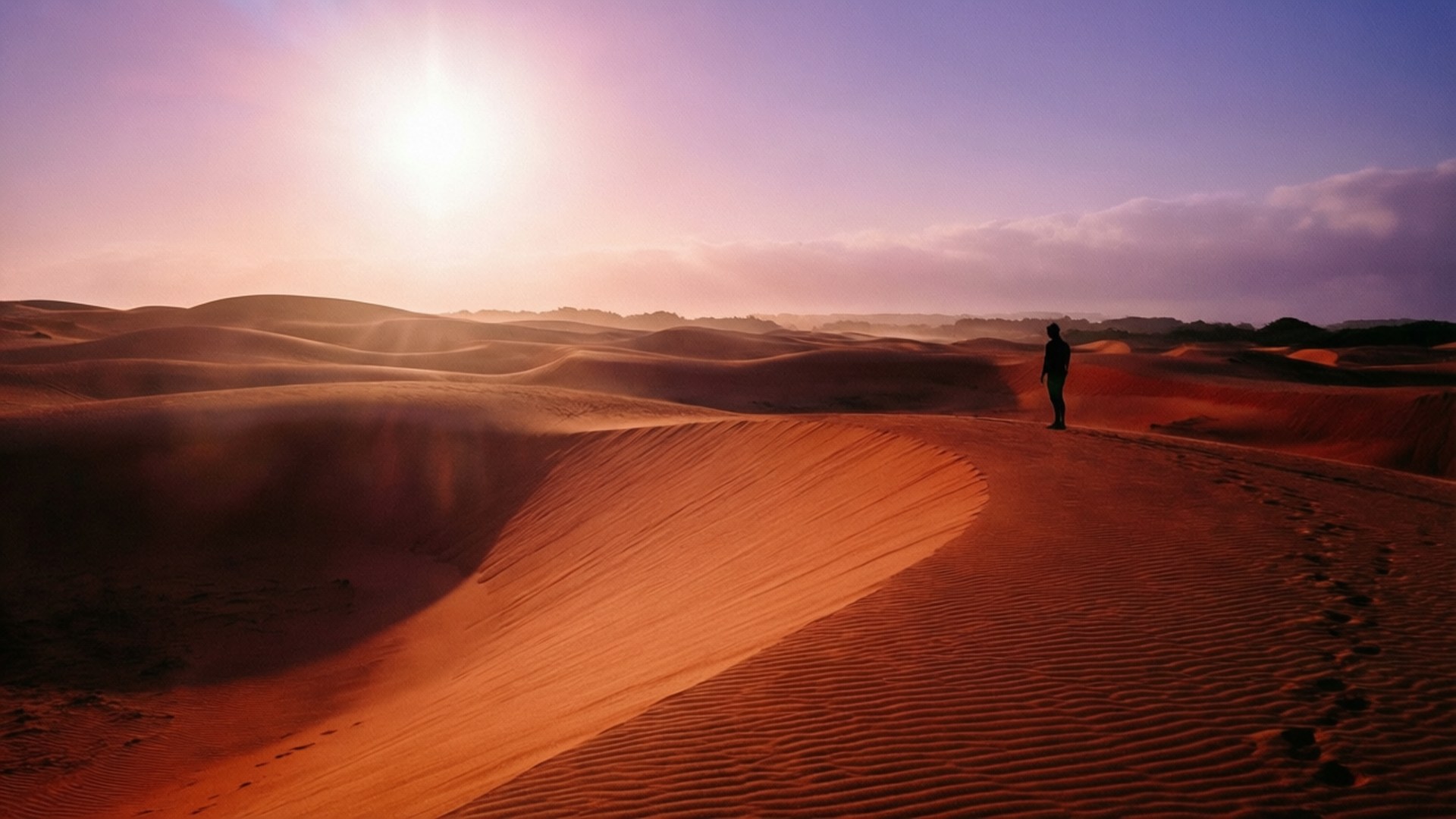 Mui Ne Sand Dunes 壁紙 — 一個孤獨的身影站在廣闊的橙色沙丘頂上，在發光的紫色日落和燦爛的陽光下。