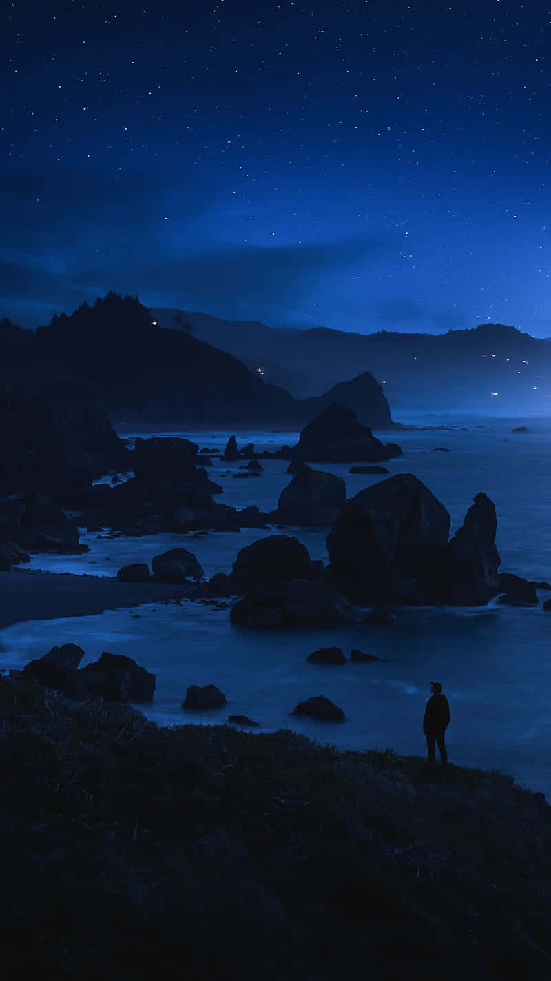  壁紙 — 一個孤獨的身影夜晚站在懸崖上，俯瞰星空和發光的海洋，以及岩石海岸。