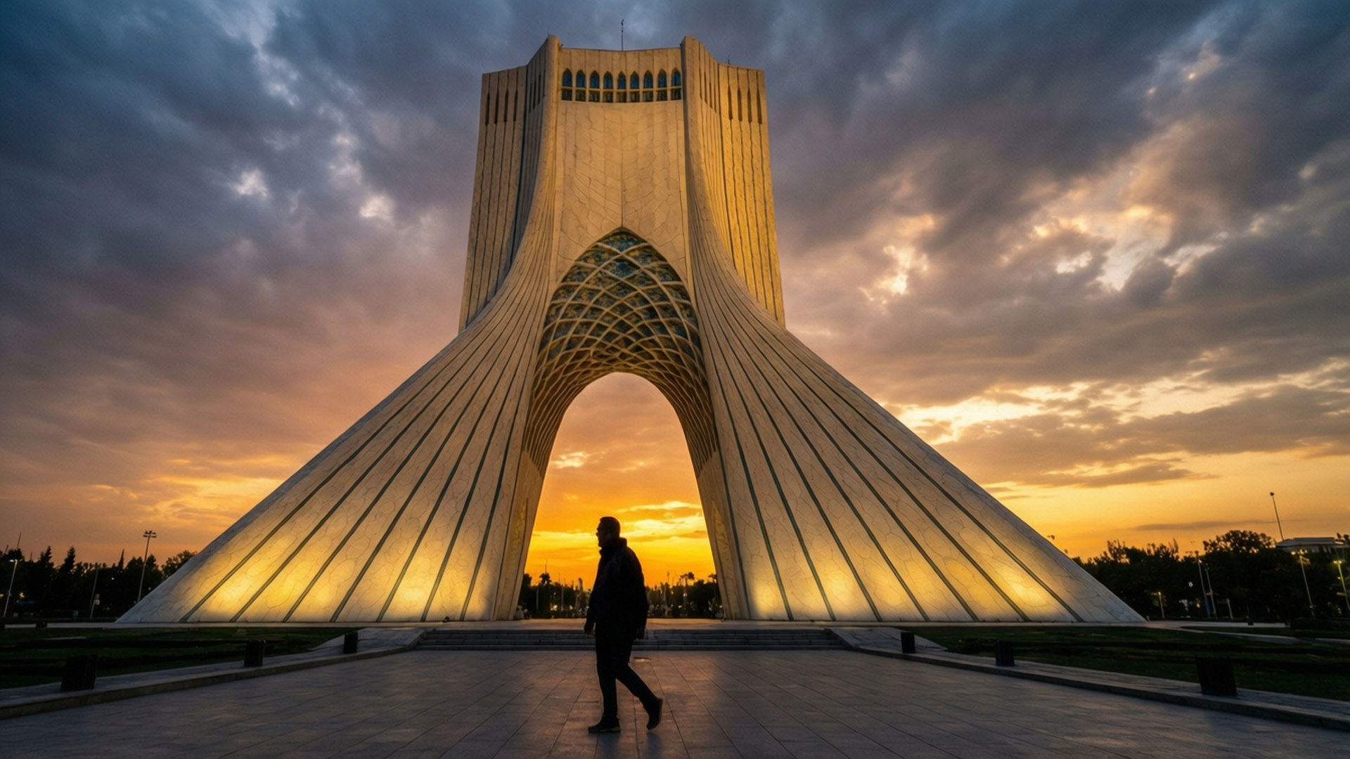 Tehran 桌布 壁紙 — 日落時分，一個孤獨的身影在燈火通明的阿扎迪塔下行走，在戲劇性的橙色和灰色雲層中投下長長的影子。