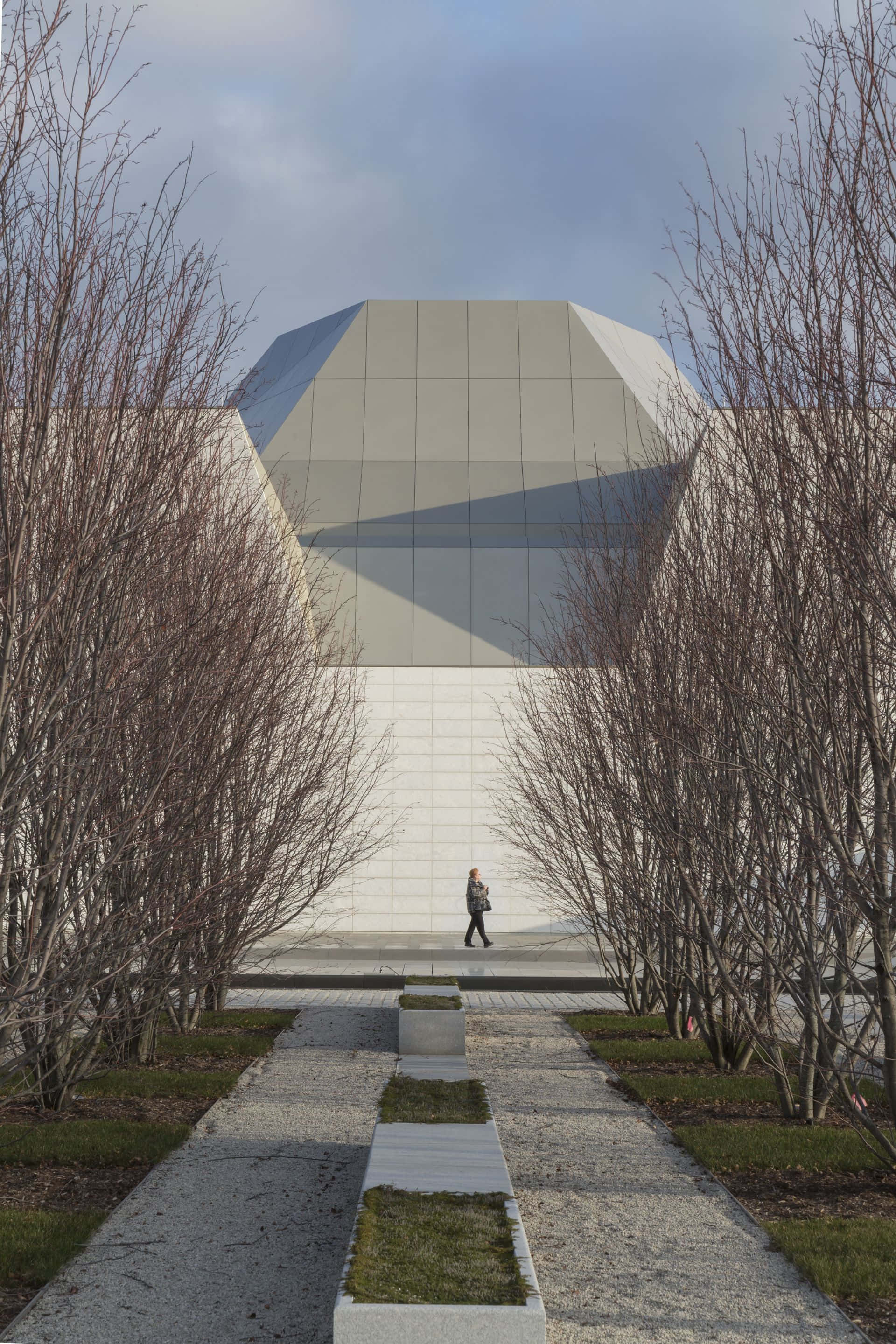 Aga Khan Museum 桌布 壁紙 — 一個孤獨的人沿著一條兩旁光禿樹木的碎石路，走向一座在陰天下引人注目的幾何現代建築。