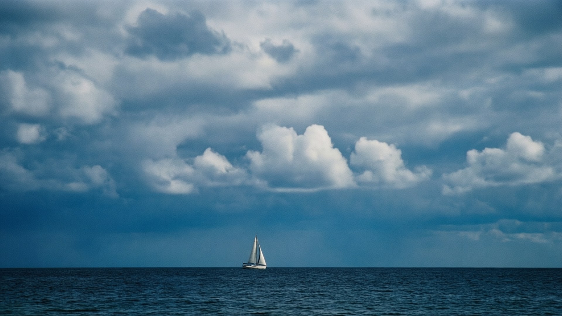 Sailboat Under Clouds 壁紙 — 一艘孤獨的白色帆船在平靜的深藍色水域中航行，下方是廣闊而戲劇性的天空，充滿了有紋理的雲彩。