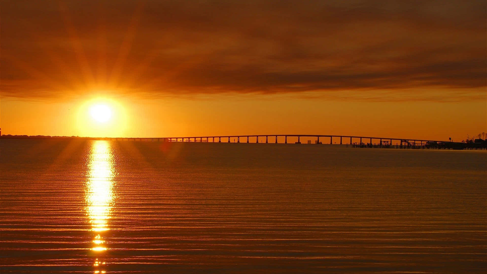 Sunset Reflection 桌布 壁紙 — 一座長橋橫跨平靜的水面，夕陽如火般橙紅，在水面上投射出一條燦爛的路徑。