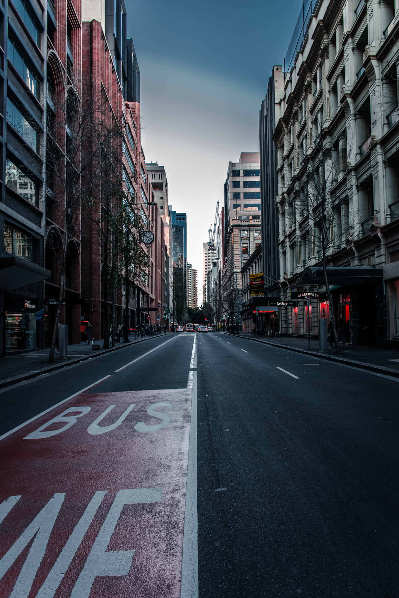  壁紙 — 一條長長的空蕩蕩的城市街道，兩旁是高樓大廈，在陰鬱的暮色天空下，帶有紅色公車專用道標記。