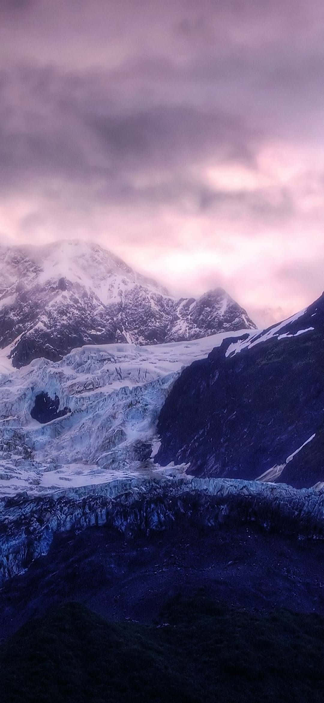 一條冰川在紫色暮色天空下蜿蜒穿過積雪覆蓋的山脈。 — iPhone 13 Pro 壁紙