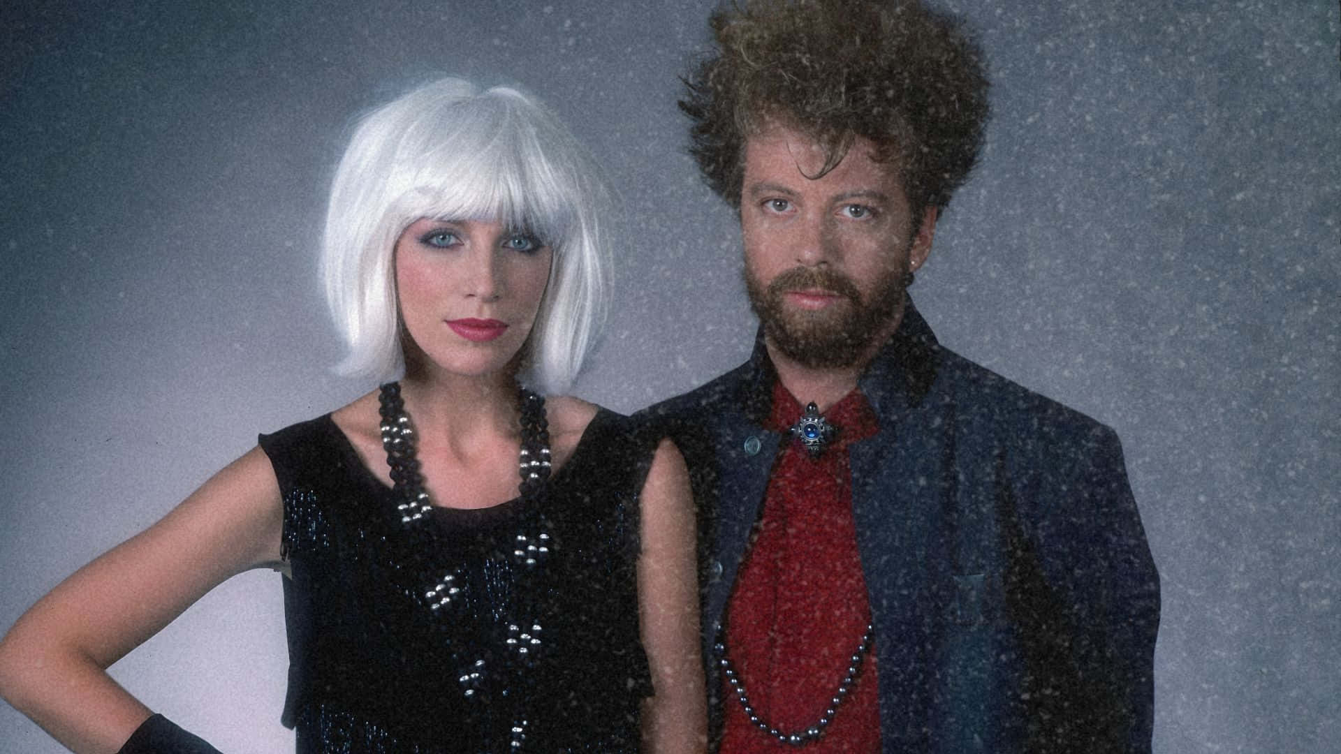  壁紙 — 一男一女身穿1980年代時尚服飾，在雪白的灰色背景前擺姿勢，燈光效果戲劇化。