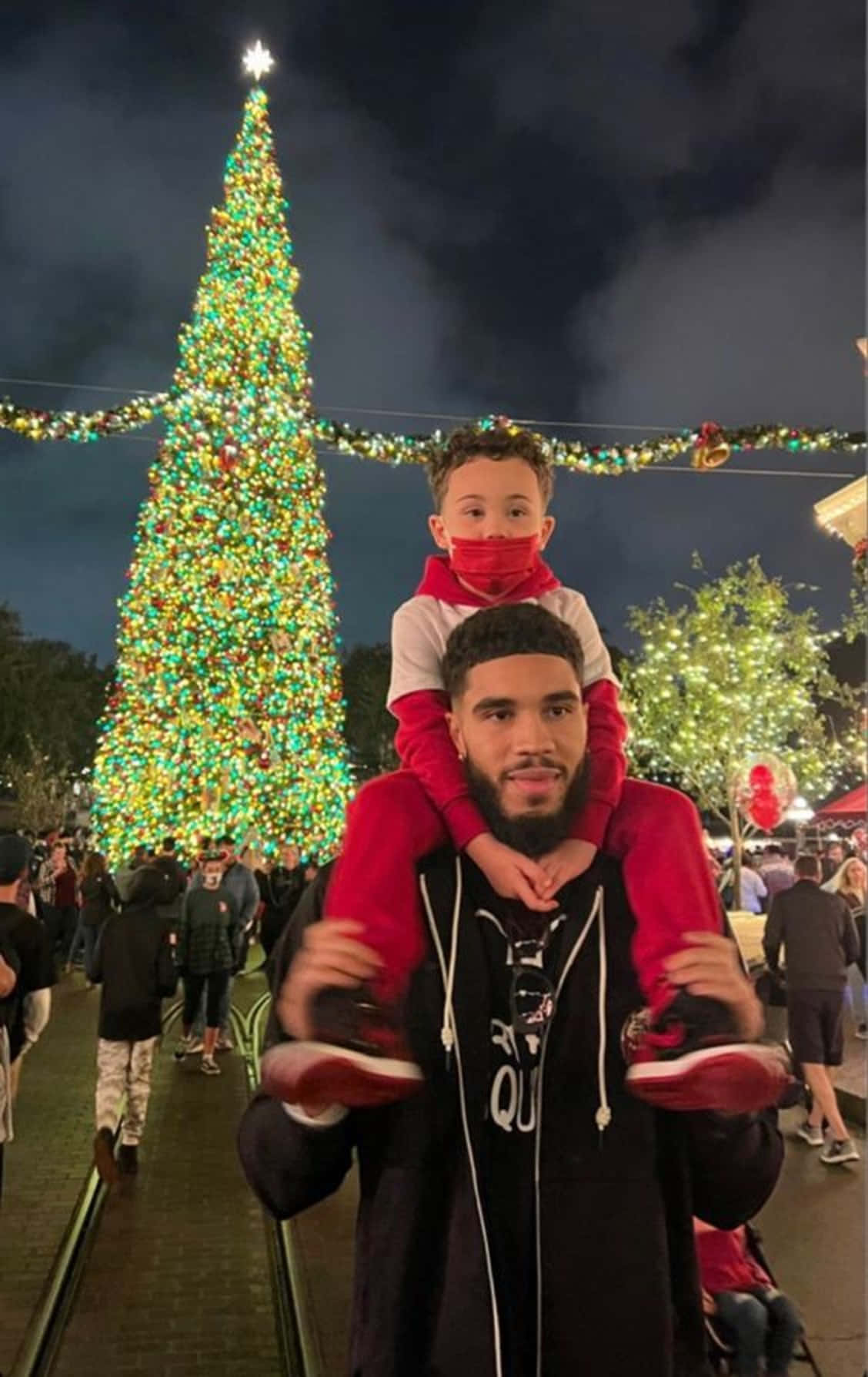 父親在夜晚於發光的聖誕樹附近將孩子扛在肩上。 — NBA聖誕節 壁紙