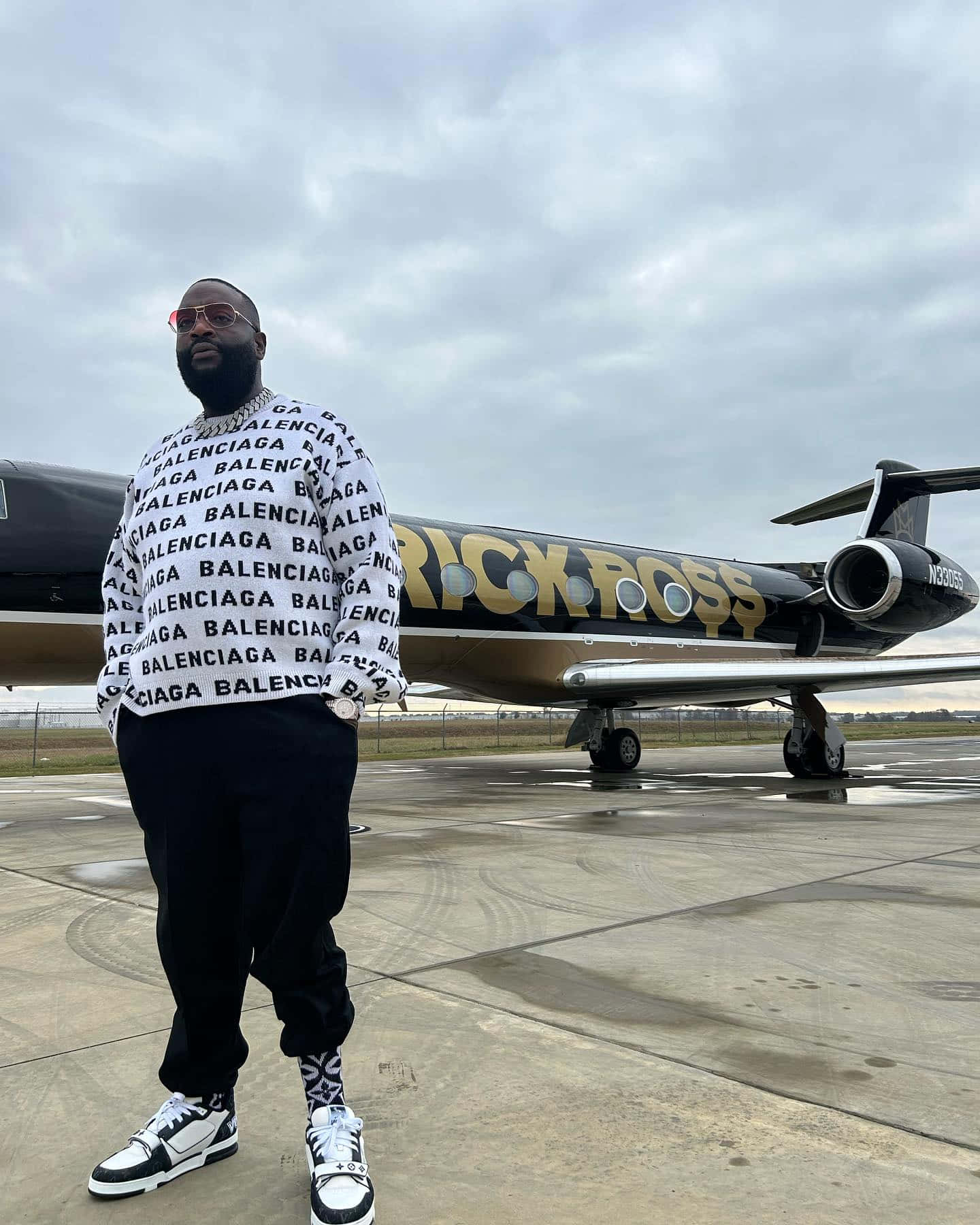  壁紙 — 一名穿著 Balenciaga 衛衣的男子自信地站在機場停機坪上，旁邊是一架印有金色 'RICK ROSS' 字樣的私人飛機。