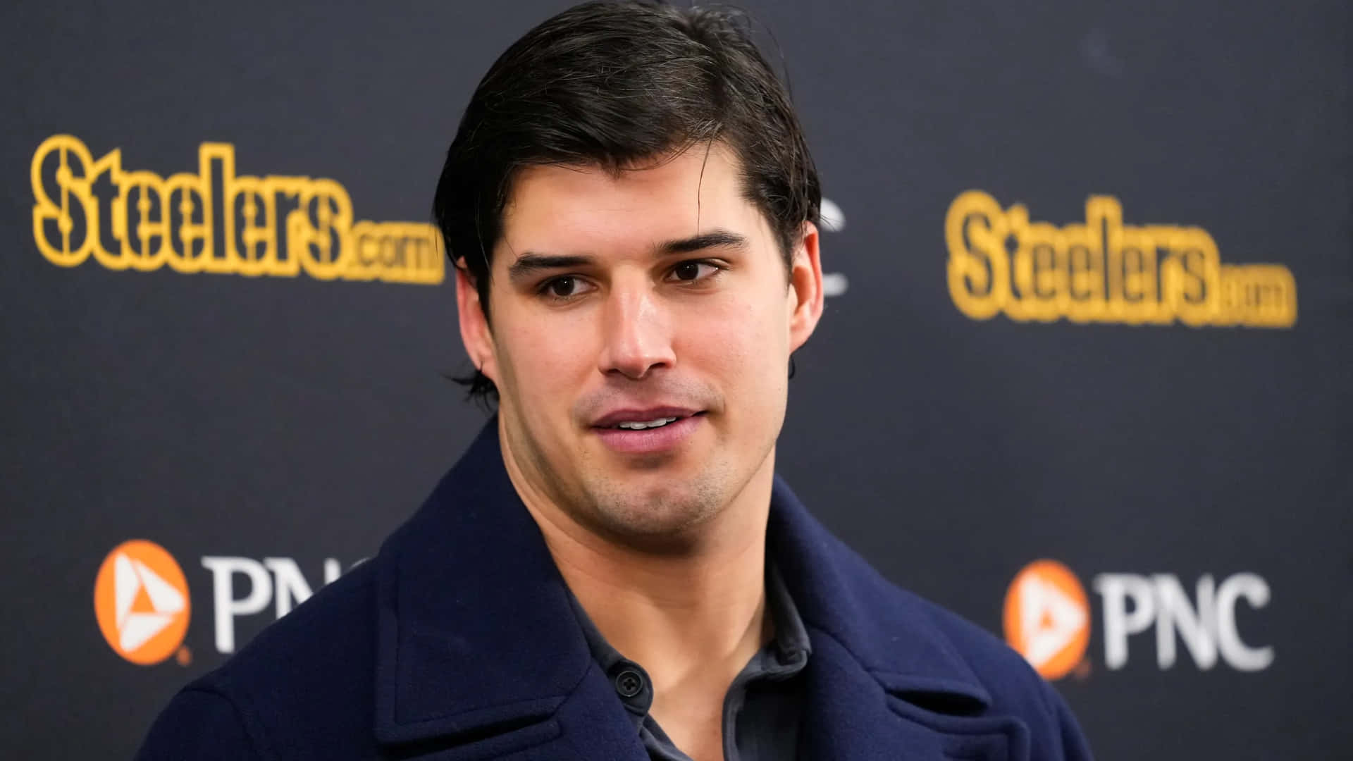  壁紙 — 一名身穿海軍藍外套的男子在Steelers.com品牌背景前對著鏡頭微笑。