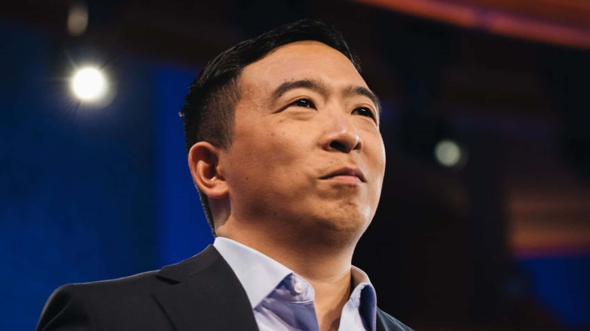 政治 桌布 壁紙 — 一名身穿西裝的男子在明亮的舞台燈光下，帶著沉思的表情向上仰望。
