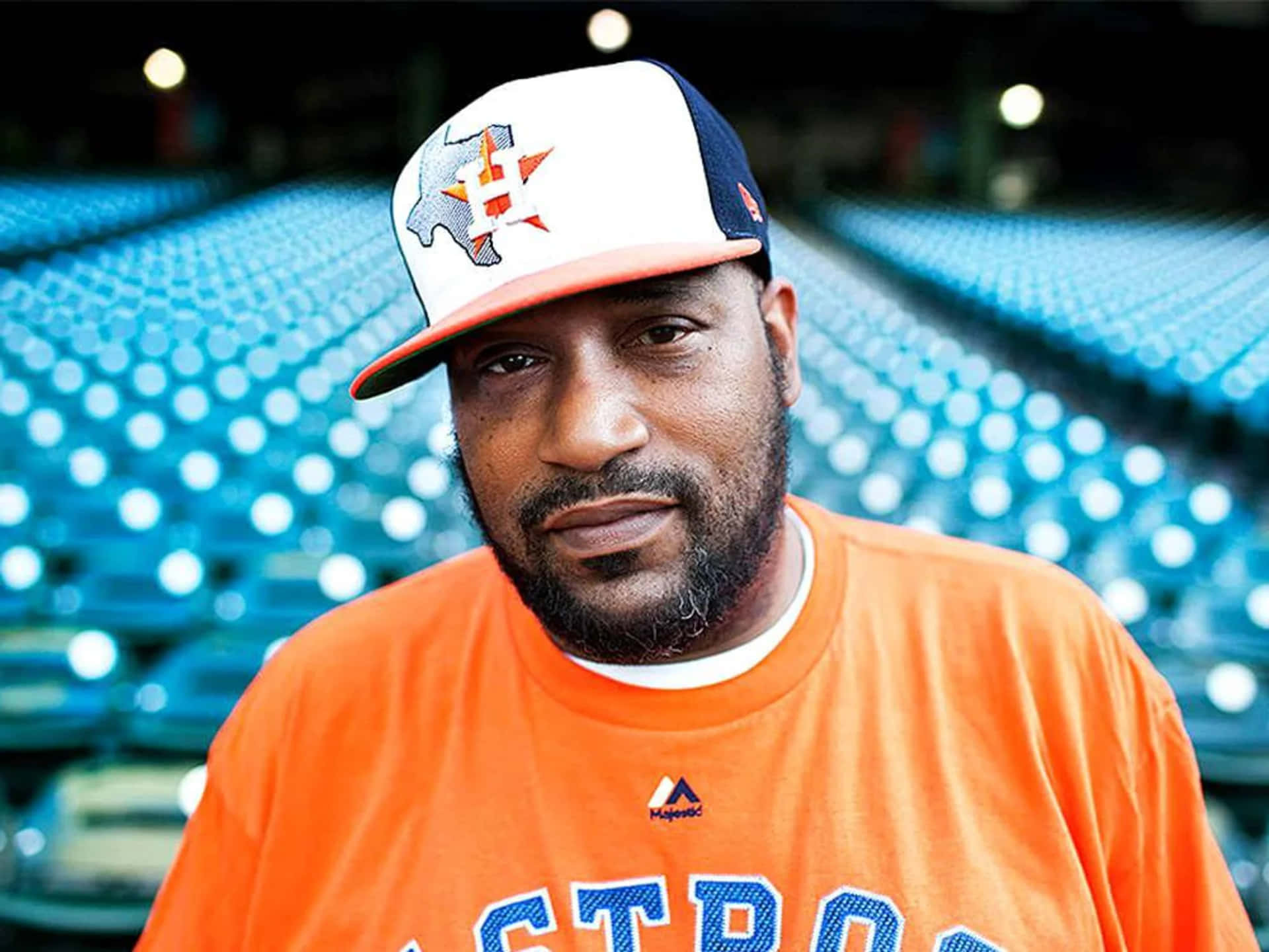  壁紙 — 一名身穿橙色Astros T恤和球隊帽子的男子，在模糊的藍色體育場座位背景下直視鏡頭。