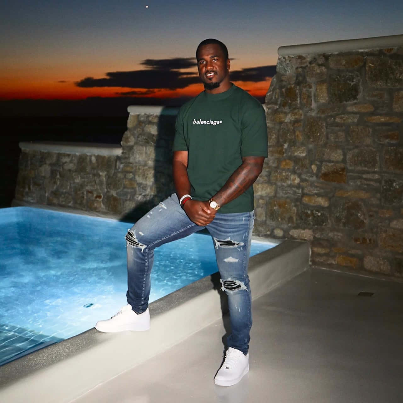  壁紙 — 一個穿著破洞牛仔褲和綠色Balenciaga T恤的男人在日落時分隨意地靠在發光的游泳池旁。
