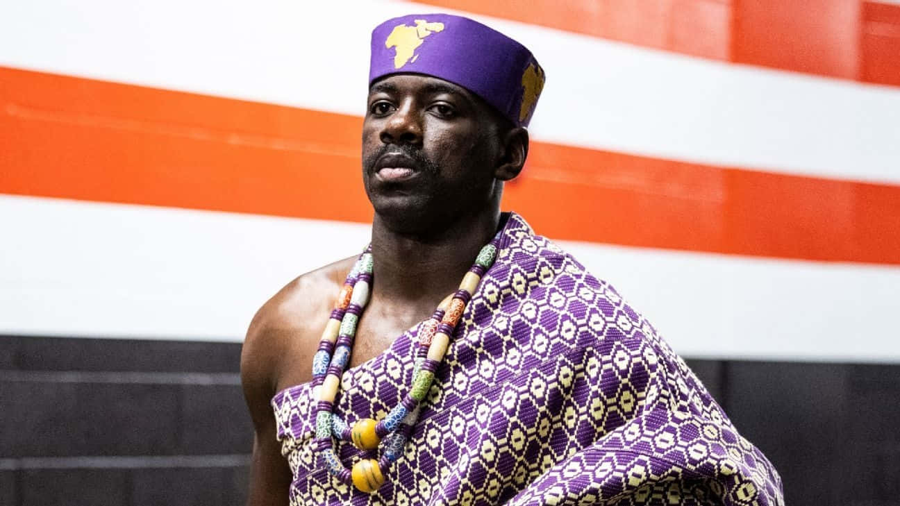  壁紙 — 一名身穿傳統紫色和金色非洲服飾的男子，自信地站在大膽的橙色和白色條紋牆前。