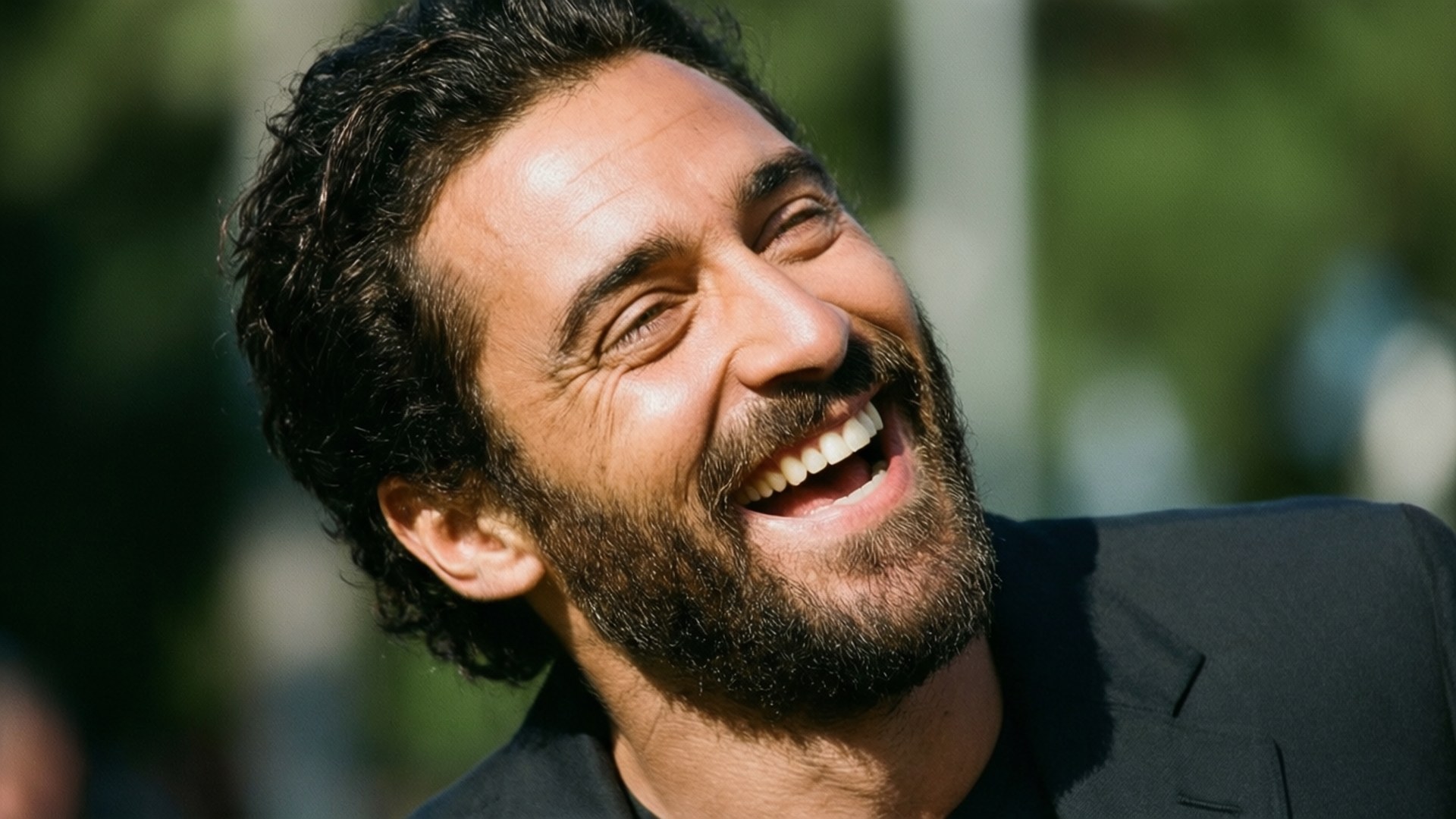 Joyful Laughter 壁紙 — 一個擁有深色捲髮和鬍鬚的男人在明亮的陽光下穿著黑色西裝在戶外開懷大笑。