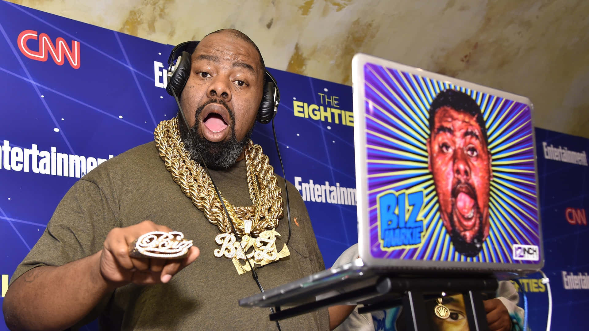  壁紙 — 一名戴著耳機和金鏈的男子手持「Biz Markie」餅乾，而筆記型電腦在充滿活力的太陽爆發背景上顯示他驚訝的表情。