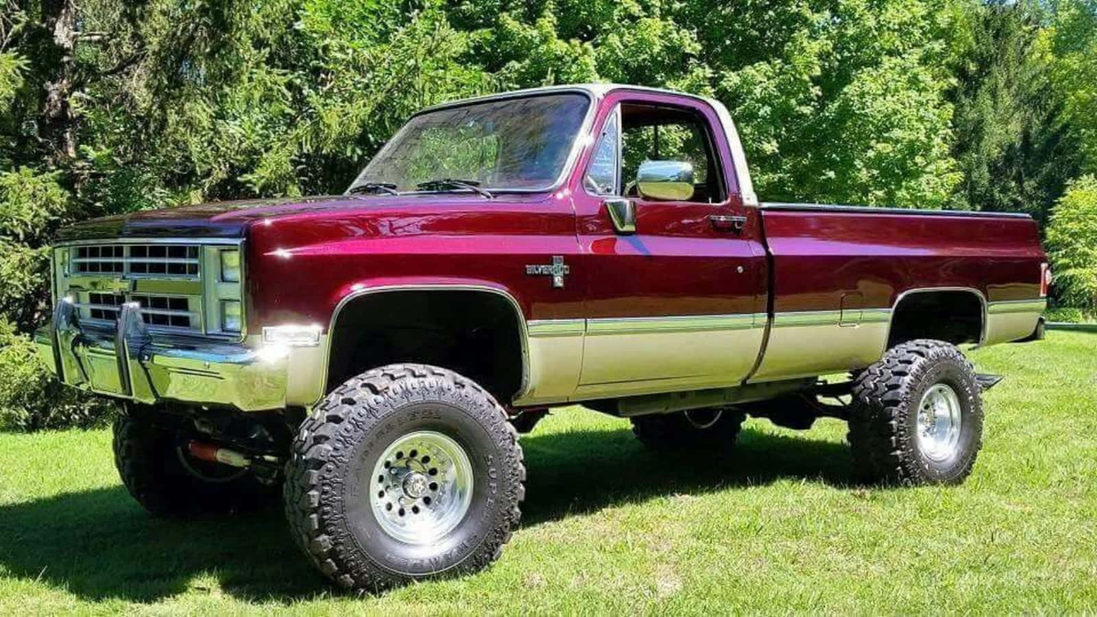Square Body Truck 桌布 壁紙 — 一輛栗色升高版雪佛蘭K10皮卡車，配備鍍鉻飾件和特大輪胎，驕傲地停放在陽光明媚的草地上，四周環繞著茂密的綠樹。
