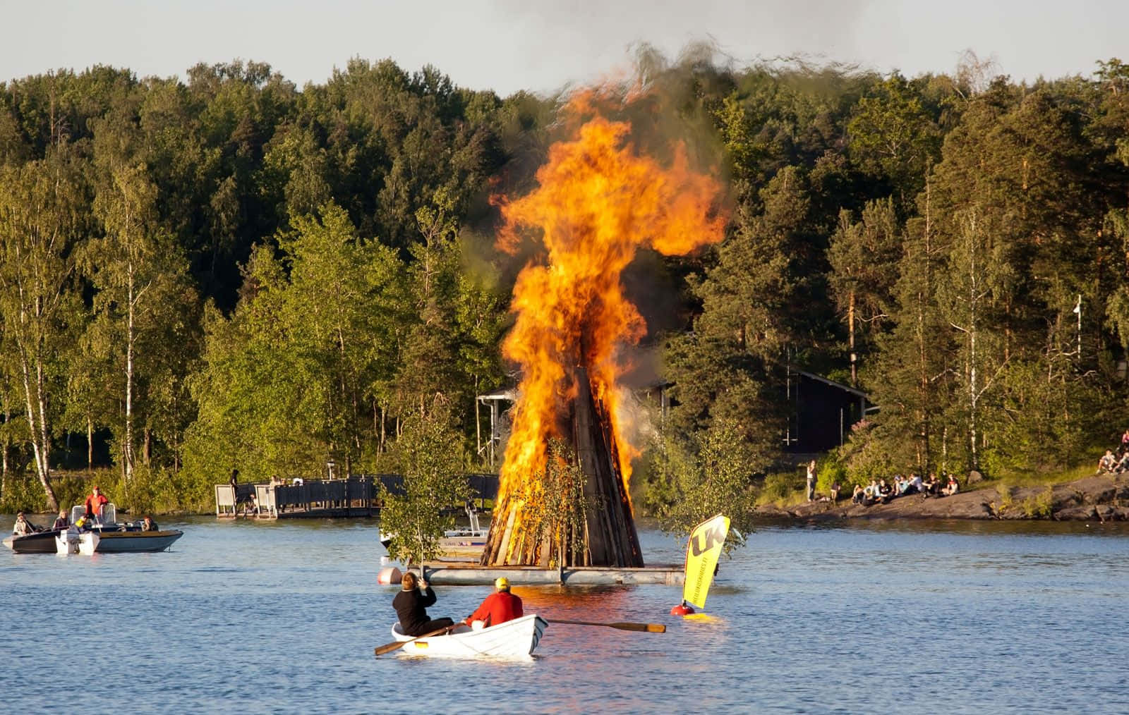  壁紙 — 一個巨大的營火在湖上明亮地燃燒，人們在船上觀看並在附近划船，周圍是茂密的綠色森林。