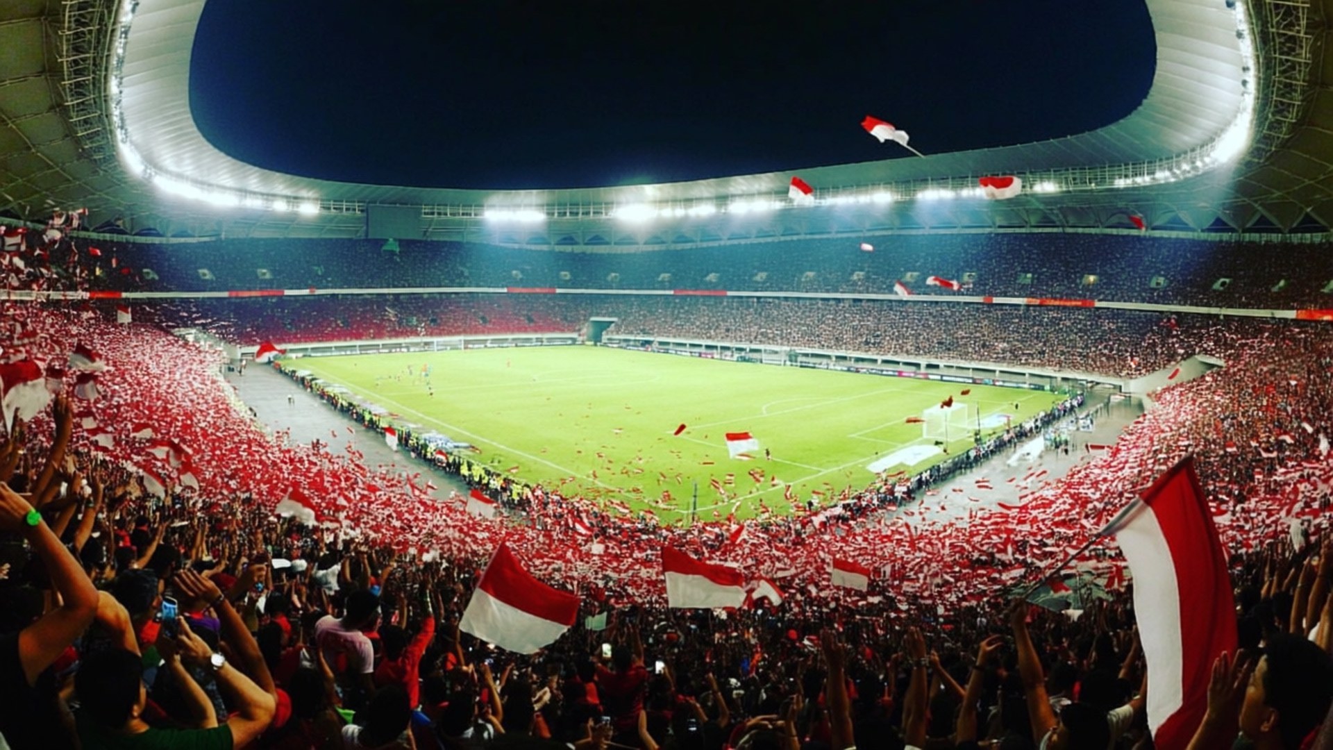 GBK Stadium Roar 壁紙 — 夜間一個巨大的體育場，充滿了歡呼的球迷，在充滿活力的綠色足球場上揮舞著紅白旗幟。