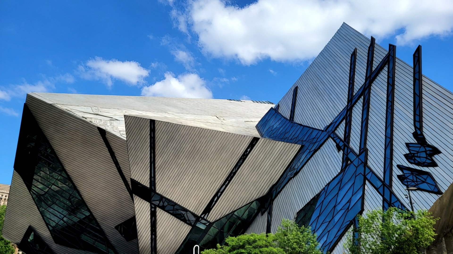 ROM Crystal Facade 壁紙 — 一座現代角形建築，採用玻璃和金屬面板，在綠色樹木的背景下反射藍天和白雲。