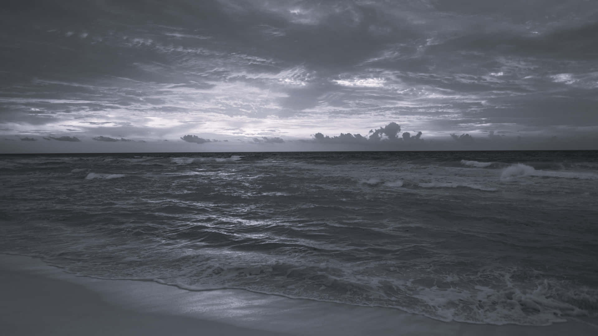  壁紙 — 一幅陰鬱的黑白海景，翻騰的海浪在厚重的烏雲天空下拍打著沙灘。