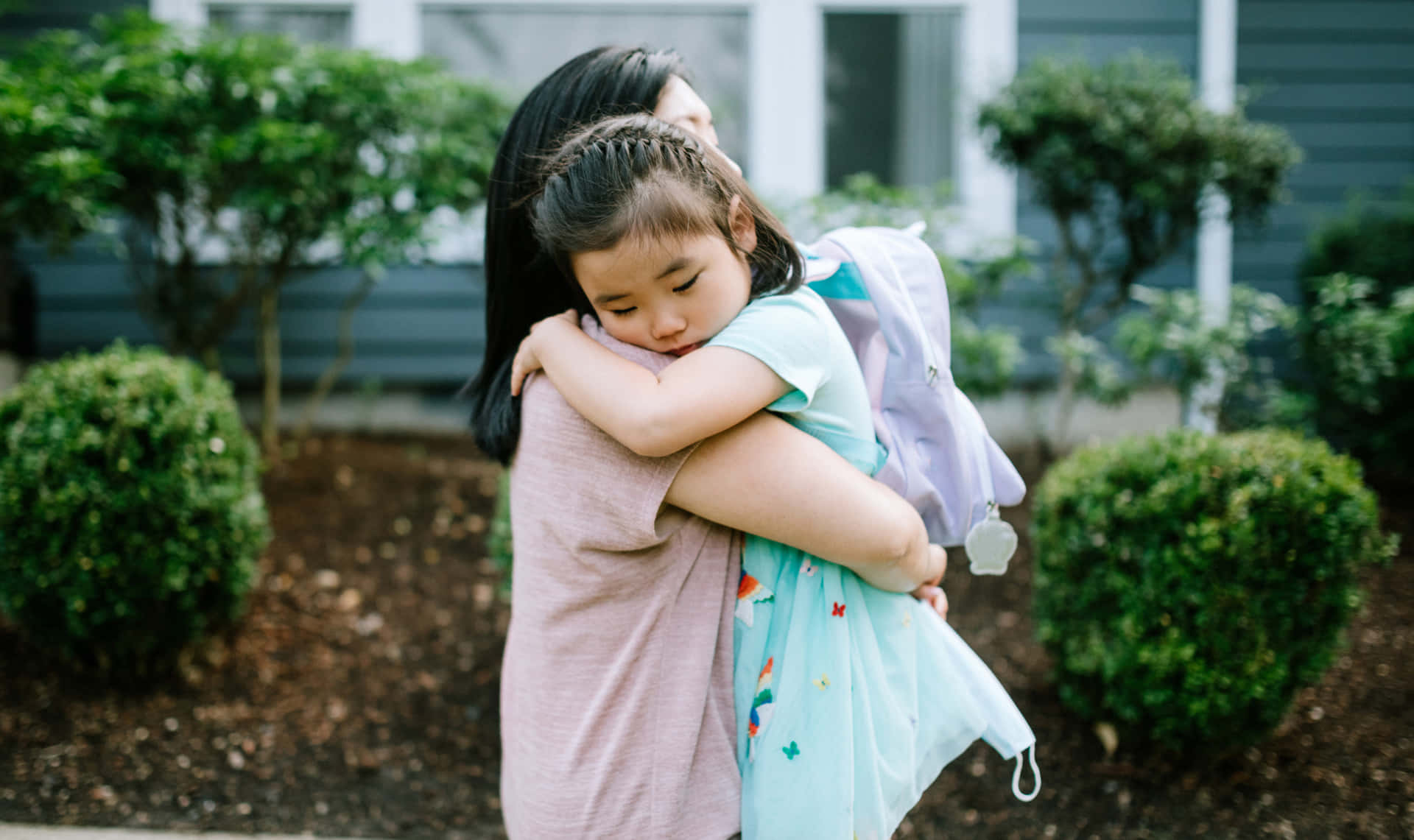  壁紙 — 一位母親在屋外的花園裡擁抱她穿著背包的小女兒。