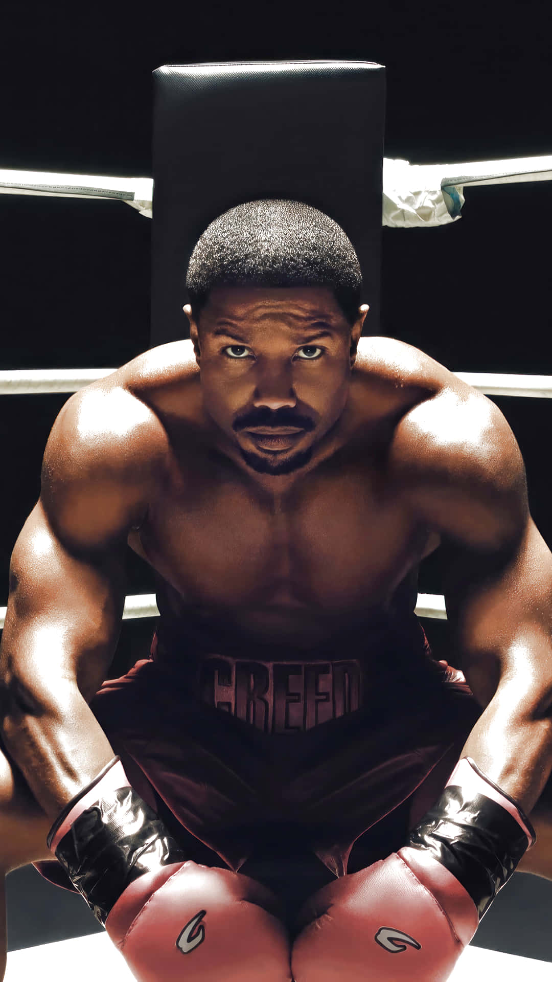  壁紙 — 一位肌肉發達的拳擊手戴著紅色手套和穿著「CREED」短褲，在燈光昏暗的擂台上激烈地蹲伏，直視鏡頭。