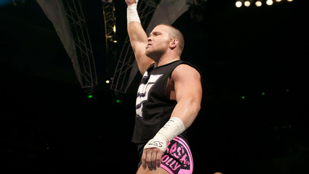 Crash Holly 桌布 壁紙 — 一位肌肉發達的摔角手身穿黑色和粉紅色裝備，在明亮的場館燈光下勝利地舉起手臂。