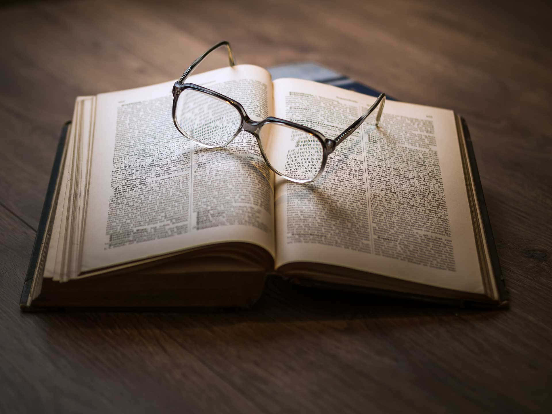 Dark Book Aesthetic 桌布 壁紙 — 一副眼鏡放在一張深色木桌上打開的字典上。