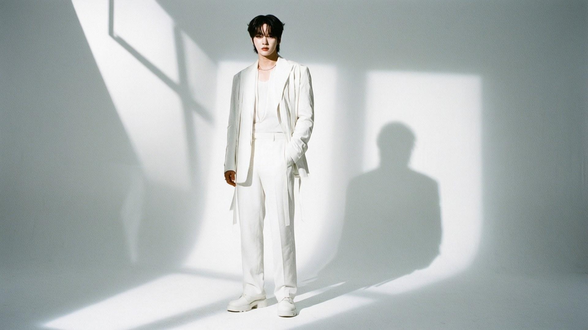 White Minimalism 壁紙 — 一個穿著白色西裝的人站在光線明亮的攝影棚中，背後牆上投射出銳利的幾何陰影。