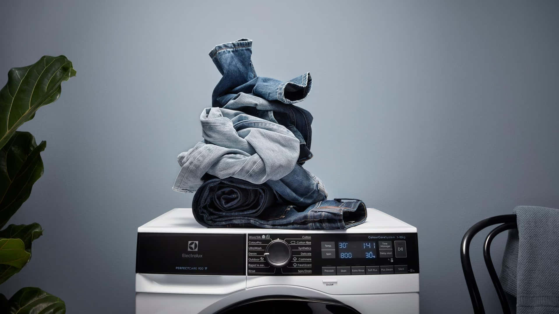  壁紙 — 一堆牛仔褲和灰色襯衫放在現代白色 Electrolux 洗衣機上，背景是柔和的灰色牆壁。