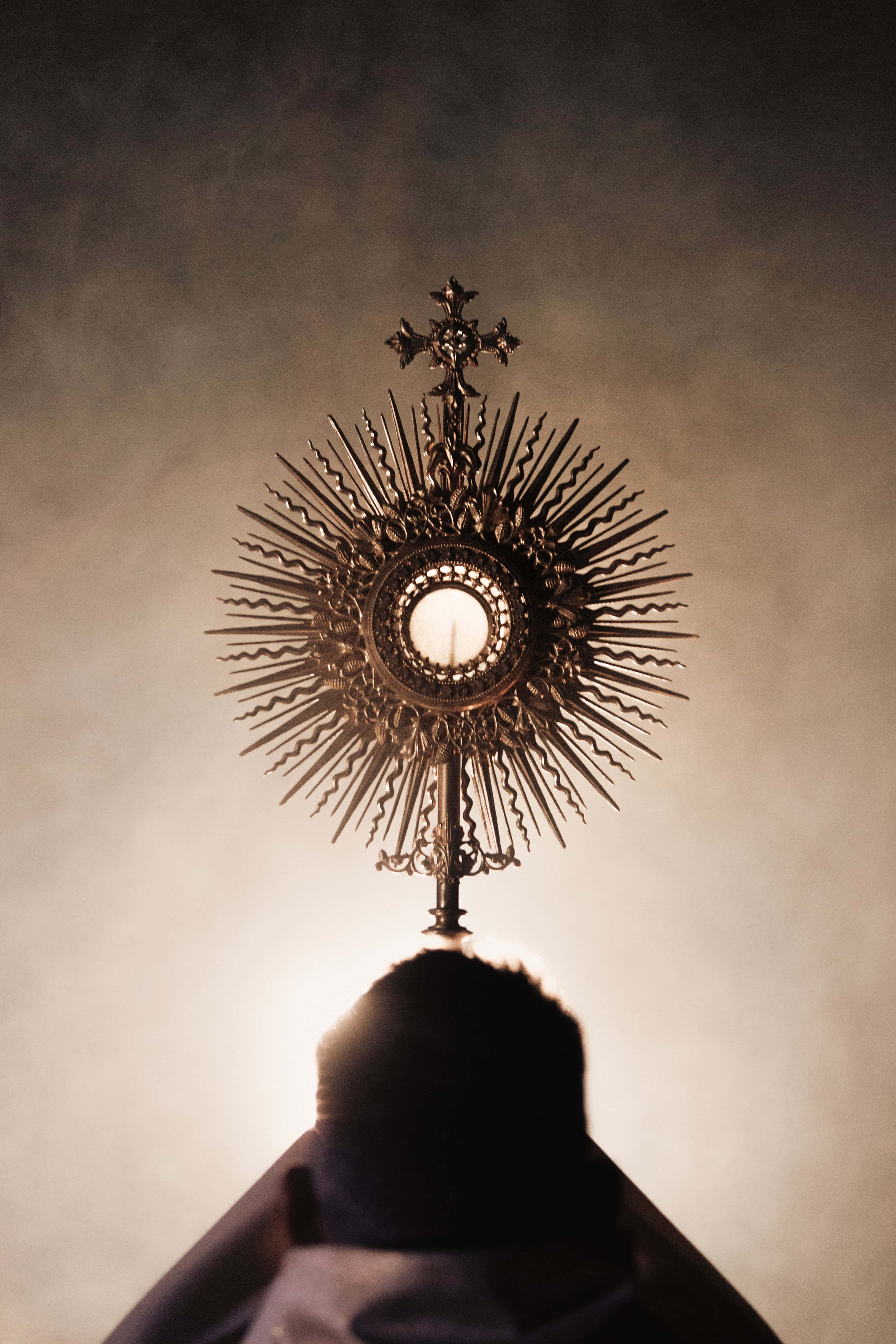 Catholic 桌布 壁紙 — 一位神父以剪影形式跪在一個光芒四射的聖體光座前，聖體光座在朦朧背景下散發著神聖的光芒。