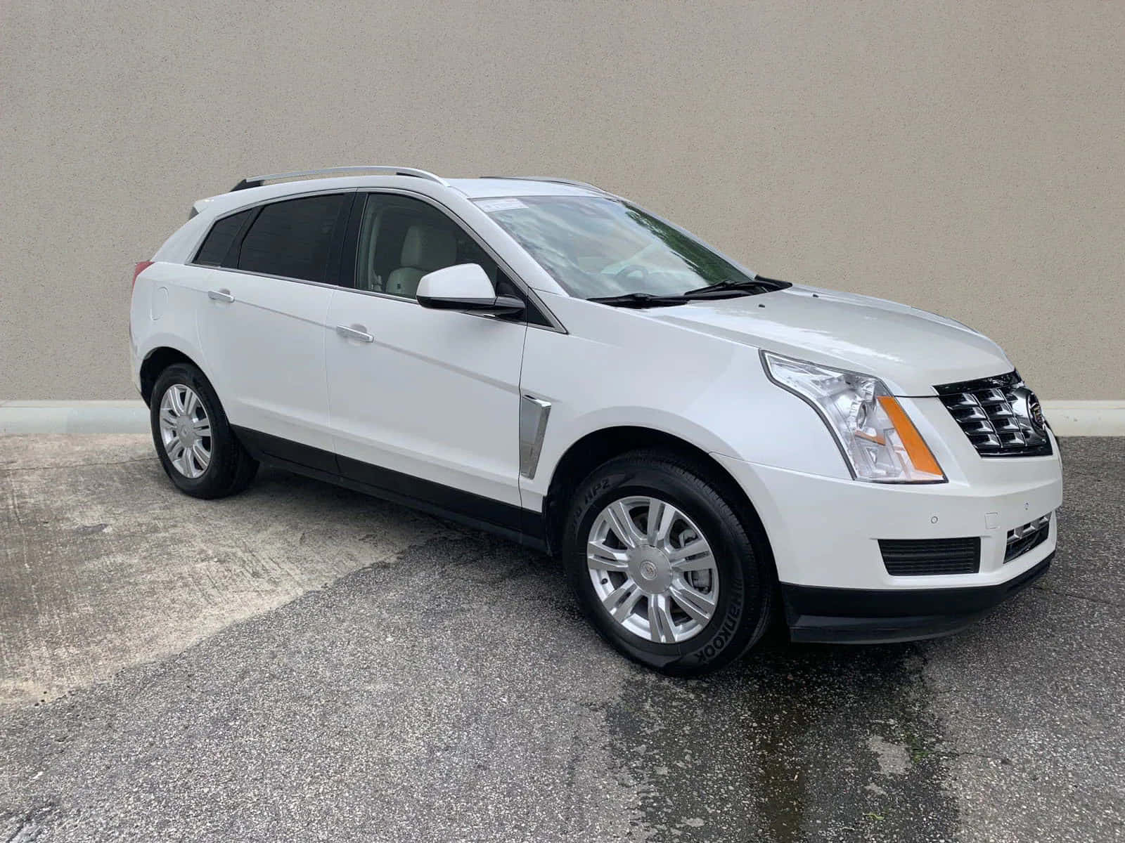 Cadillac Srx 桌布 壁紙 — 一輛純白色的Cadillac SRX SUV停放在濕漉漉的柏油路上，背景是樸素的米色牆壁，天空多雲。