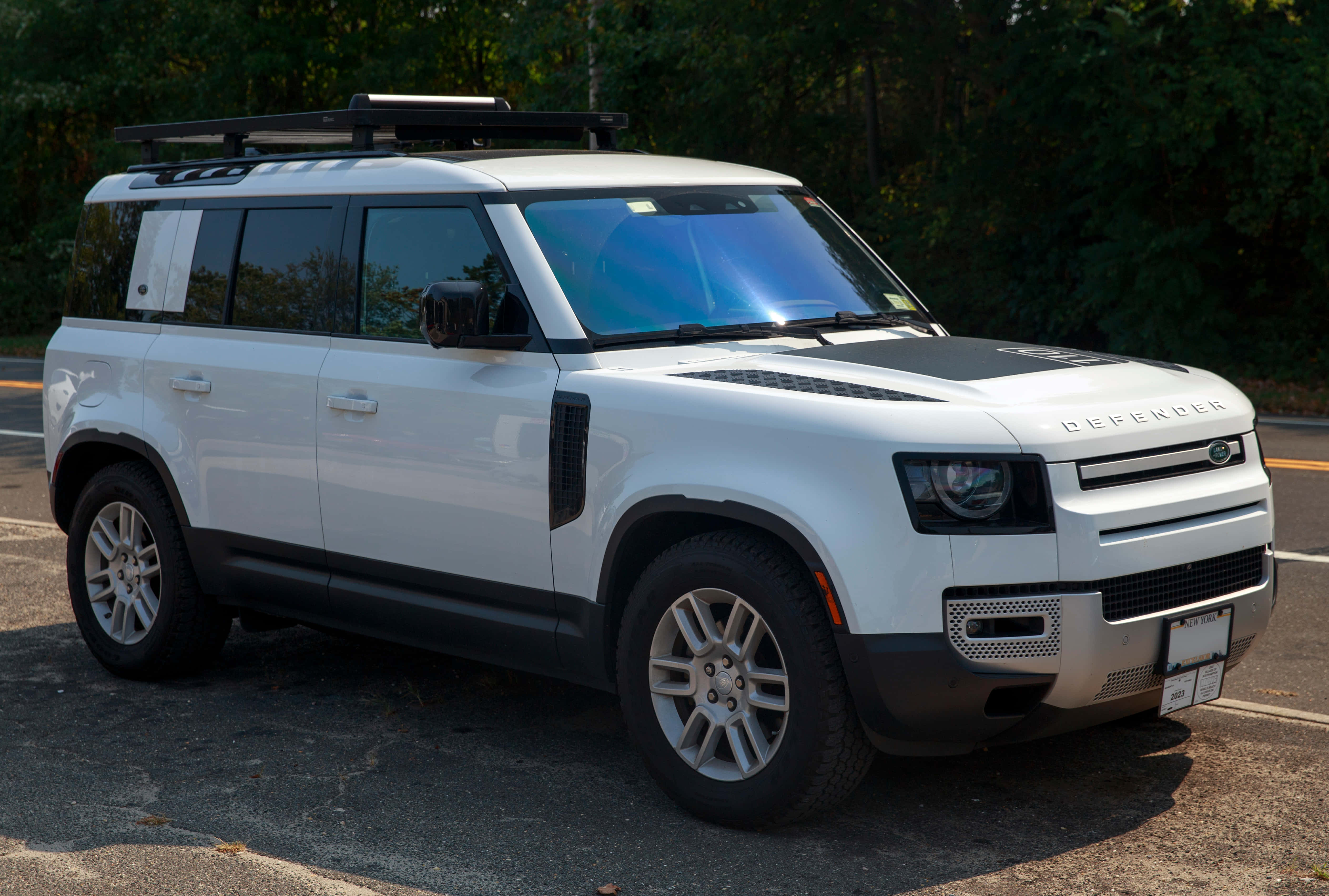 Land Rover Defender 桌布 壁紙 — 一輛純白色的 Land Rover Defender 停在柏油路上，背景是茂密的綠樹，在明亮的日光下。