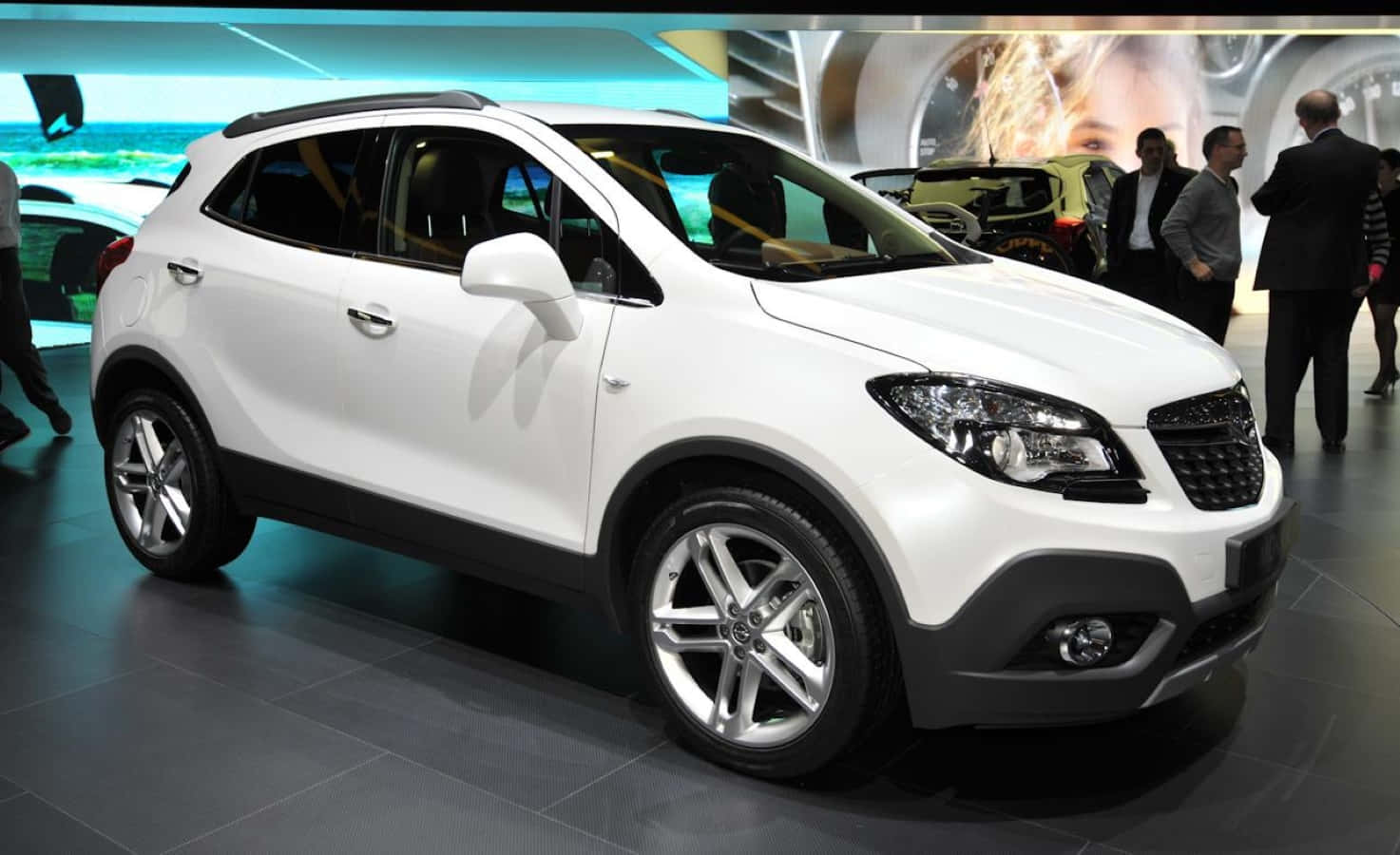 Opel Mokka 桌布 壁紙 — 一輛純白色SUV在明亮燈光下閃耀，於繁忙的車展中，與會者在附近觀看。