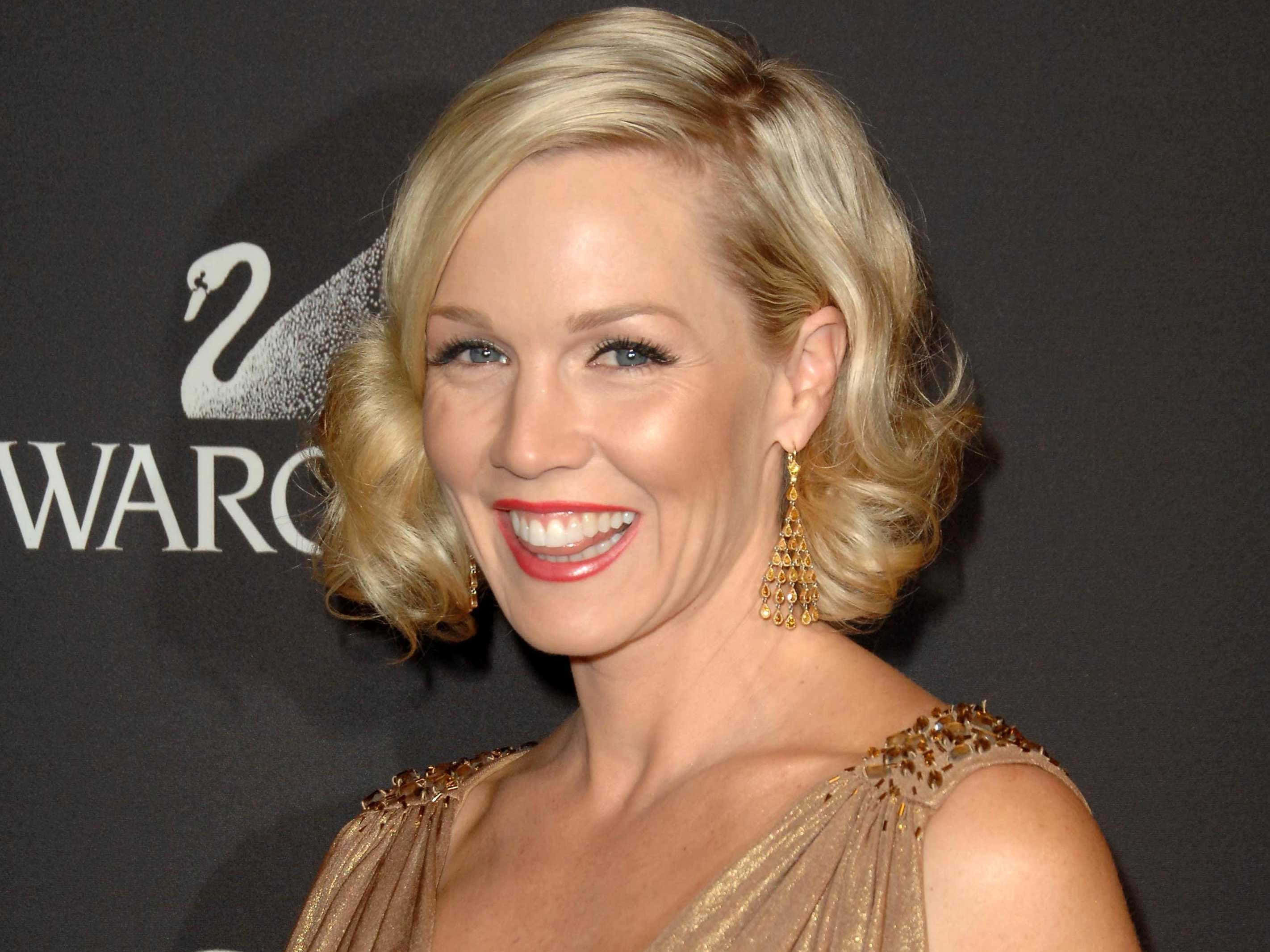 Jennie Garth 桌布 壁紙 — 一位光彩奪目的金髮女性，塗著紅色唇膏，戴著金色耳環，穿著閃閃發光的金色連衣裙，在施華洛世奇頒獎典禮上微笑。