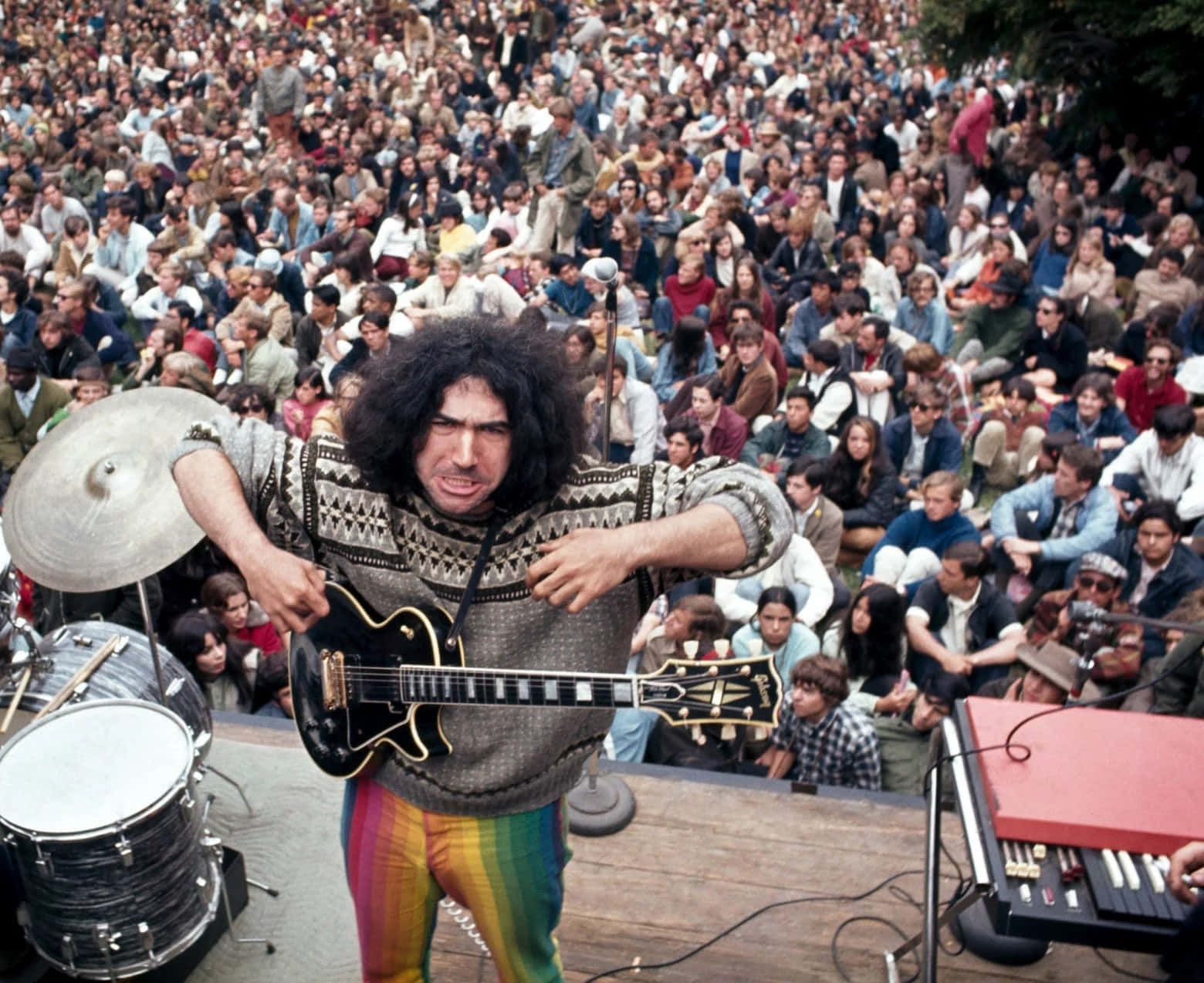  壁紙 — 一位搖滾音樂家，頭髮狂野，穿著彩虹褲，在1970年代戶外音樂會上，在大量人群面前彈奏吉他。