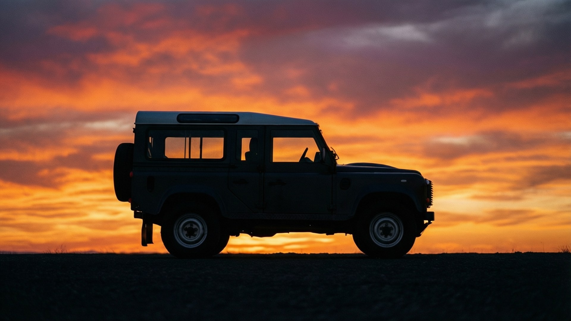 Silhouette Sunset 壁紙 — 堅固的 Land Rover Defender 剪影矗立在火熱的橙色和紫色日落天空前。